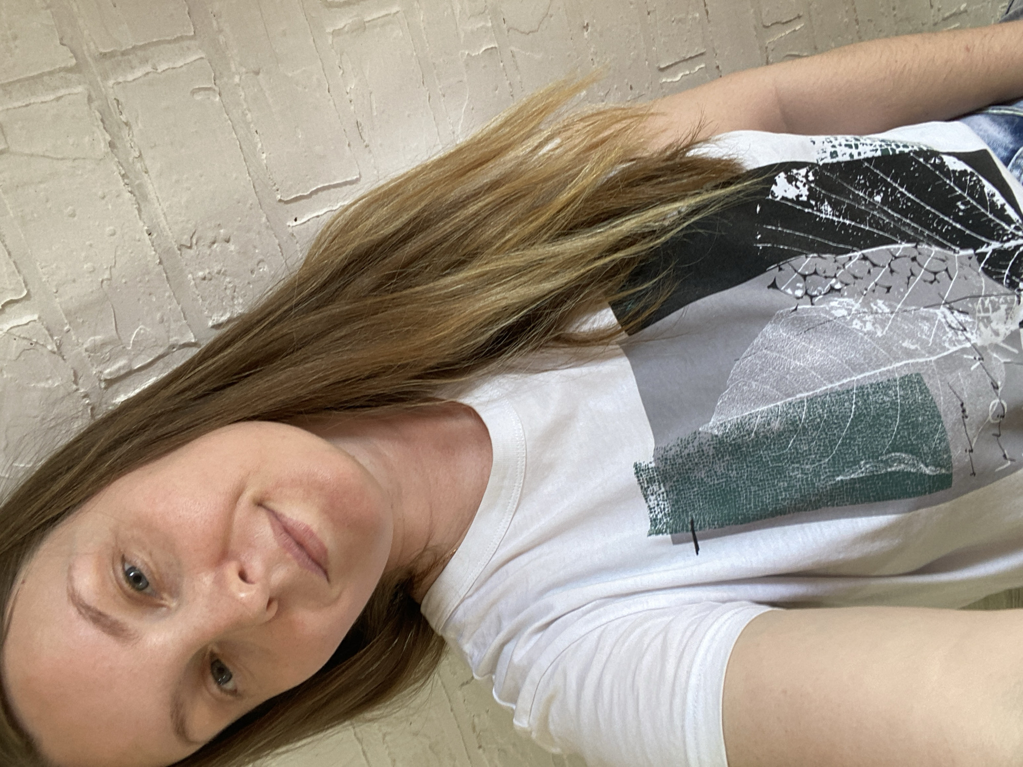 A woman is shown from the shoulders up, angled in the frame against a light brick wall. She has long brown hair with lighter highlights. She is wearing a white t-shirt with a print of leaves in shades of black, grey, and green. Her expression is neutral, and she is looking at the camera. No information indicates that the picture was taken in Noviy, Russia.