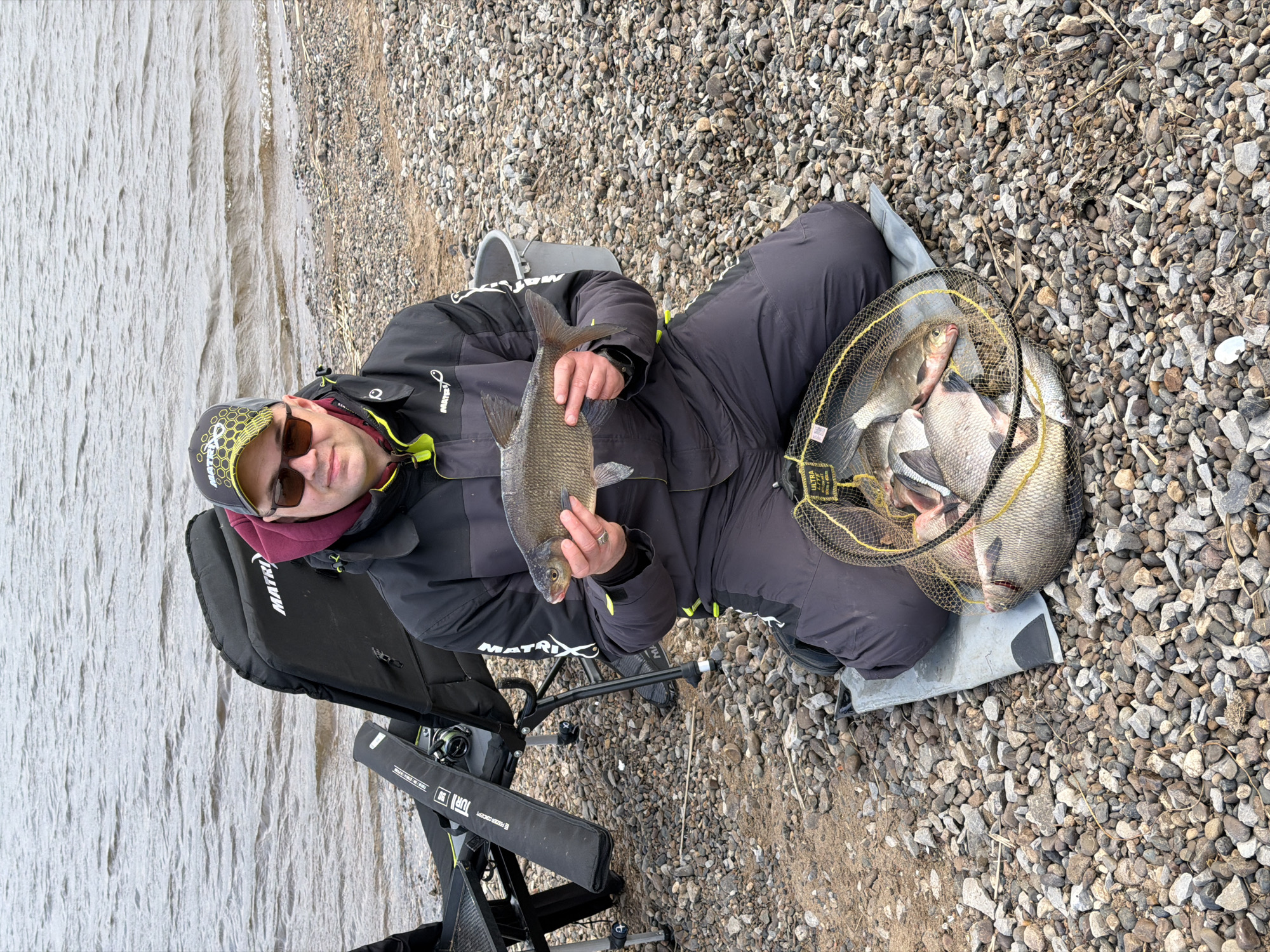 This image, taken in Chaykovsky, Russia, shows a male angler seated on a pebbly shore next to a large body of water. He is dressed in a black hooded jacket bearing a "MATRIX" logo, dark trousers, sunglasses, and a black and yellow patterned cap featuring a skull or geometric design. The man proudly holds up a large, silver-grey fish, likely a bream, with both hands.
To his right, a yellow-rimmed keepnet rests on a grey mat, filled with several other freshly caught fish of similar species. The fisherman is seated on a black fishing chair, also branded "MATRIX," which has a fishing rod and reel attached to its frame. Text like "ULTRA" and "FURY" are visible on parts of the fishing equipment.
The setting is an outdoor environment, with a shore comprised of small, light-colored pebbles and gravel. Beyond the shore lies a wide expanse of calm, light grey water, suggesting a river or large lake, consistent with the location context of Chaykovsky on the Kama River. The subtle ripples on the water and the man's warm clothing (jacket, hood, neck gaiter) imply a cool, possibly overcast, daytime atmosphere. The scene conveys a sense of accomplishment and contentment from a successful fishing trip.