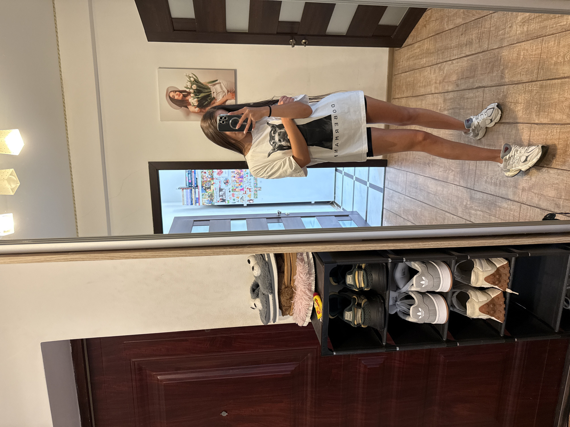 A young woman stands in a hallway, taking a mirror selfie. She has long, dark hair and is wearing a white t-shirt with a black Doberman graphic and the word "DOBERMANN" printed below it, paired with black shorts. She is also wearing white athletic shoes with grey accents. She is holding a smartphone in her right hand, angled to capture her reflection.
The hallway appears to be an apartment interior. To her left is a dark red wooden door with a single panel. Beside the door, stacked on shelves, are various shoes and slippers, including a pair of fuzzy grey slippers with cartoon eyes and several pairs of sneakers. A black shoe rack is visible.
To the woman's right, a large mirror reflects the scene. Beyond the mirror, a wooden-framed door with glass panels is open, revealing a glimpse of another room. On the wall, slightly behind and to the right of the woman, is a framed picture of a woman wearing a hat and holding a bouquet of white tulips. The floor is made of polished wooden planks laid in a diagonal pattern. Two ceiling lights with a geometric pattern are visible at the top of the frame. The overall impression is that of a residential hallway in an apartment building, likely in Chișinău, Moldova, given the context.