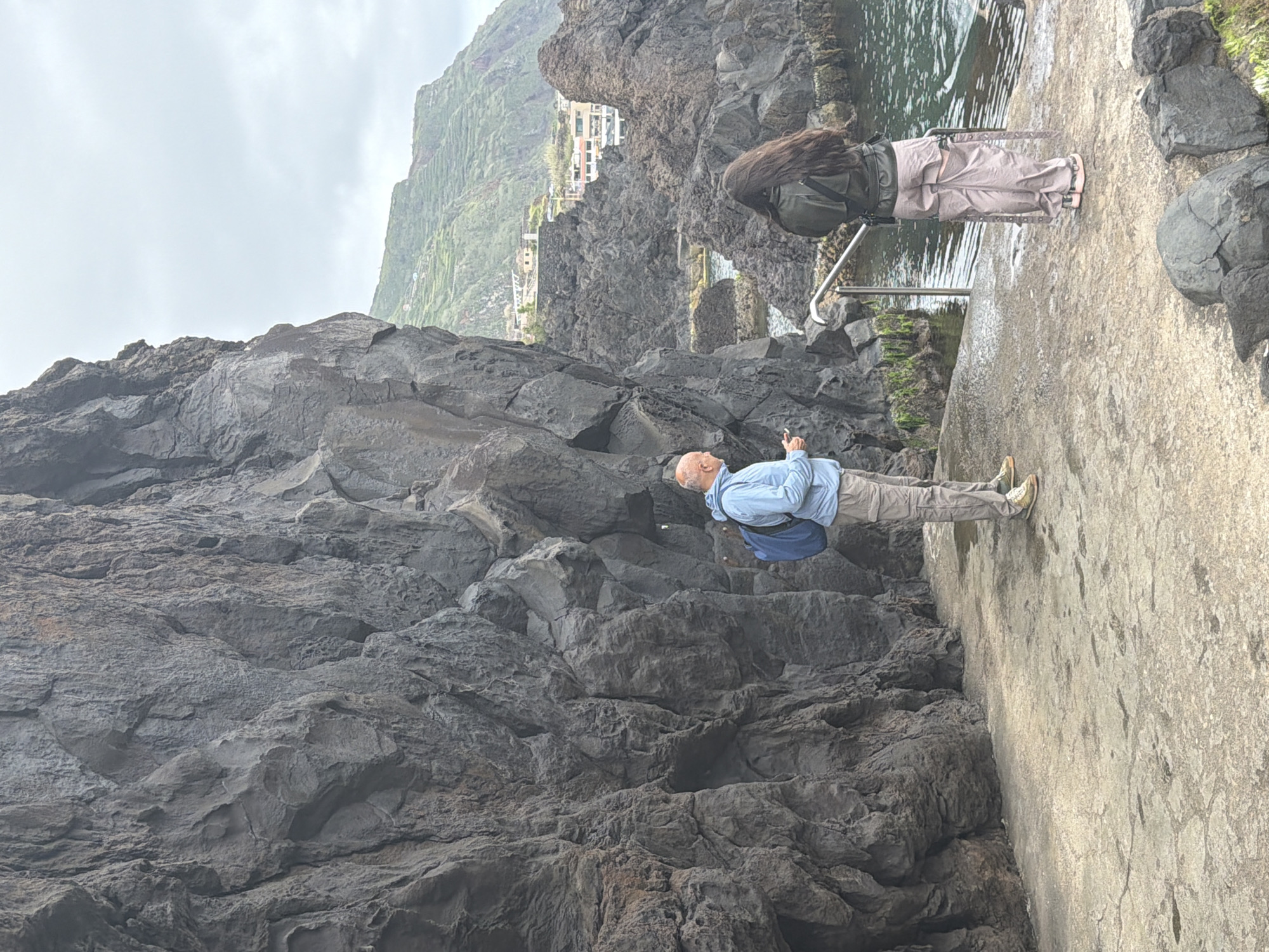 A man and woman stand on a concrete walkway bordering a volcanic rock formation and a pool of water. The man, bald with a blue shirt and tan pants, points towards the rocks. The woman, with long brown hair, wears a black jacket and light-colored pants, standing near a metal railing at the pool's edge. In the background, a cliff with buildings is visible. The scene is at the natural swimming pools of Porto Moniz, Portugal.