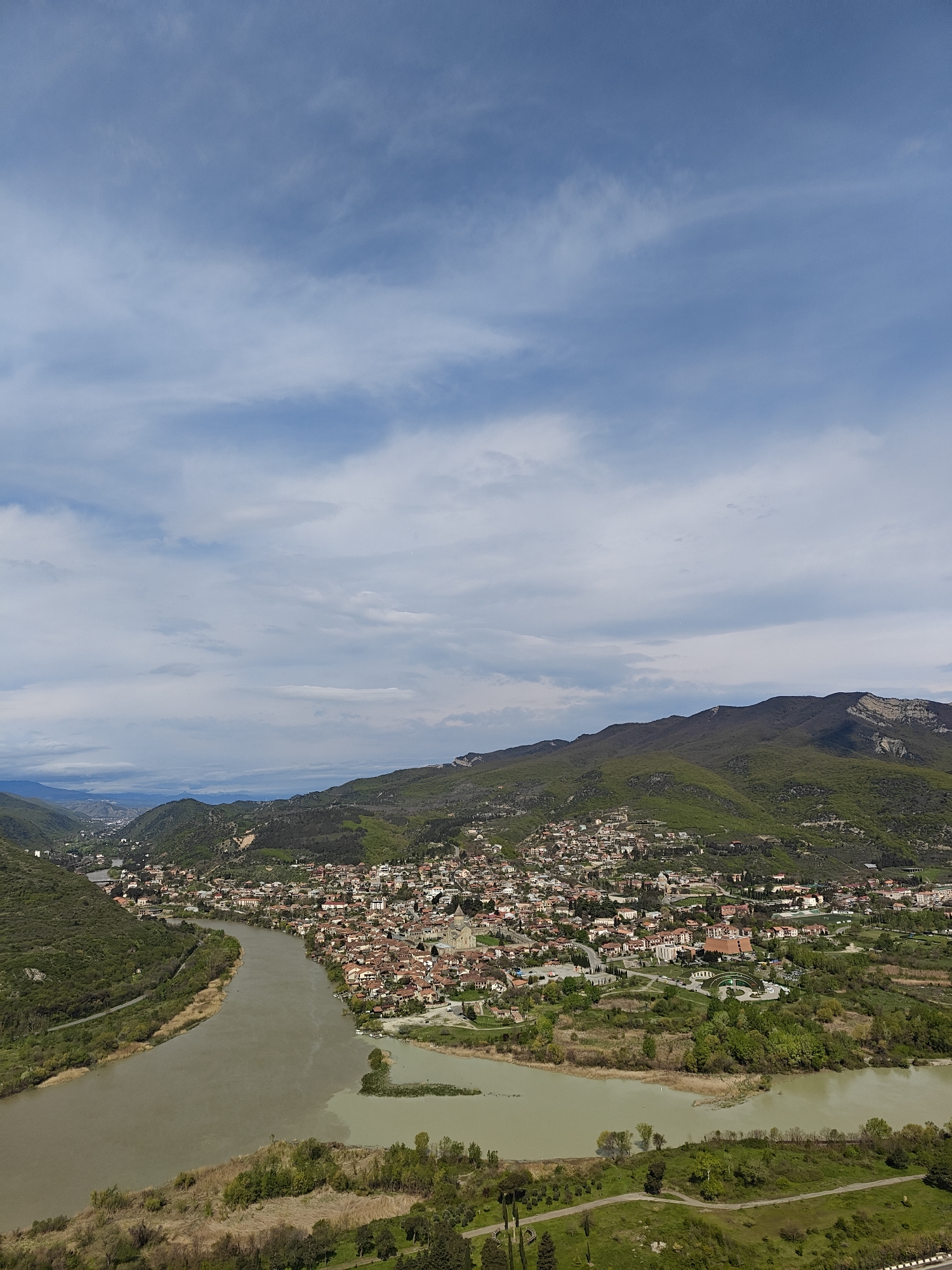 A wide-angle shot reveals a cityscape nestled at the confluence of two rivers under a cloudy sky. The buildings, primarily residential with red-tiled roofs, are densely packed. Green hills rise in the background. The rivers, differing in color and turbidity, meet in the foreground. Trees and patches of grass are visible near the riverbanks. The location is identified as Mtskheta, Georgia.
