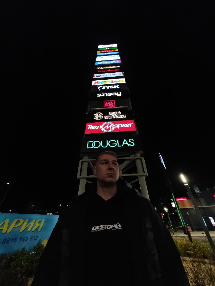 The scene depicts a male individual standing outdoors at night in Shumen, Bulgaria, against the backdrop of a prominent retail park signpost. The individual, positioned slightly left of center, is seen from the chest up, facing slightly right with a neutral expression. He wears a dark hooded sweatshirt with "DYSTOPIA / AESTHETIC HARMLESS REVOLUTIONARY" printed in white on the front.
To the right, a tall, vertical signpost displays multiple illuminated store logos stacked from bottom to top: DOUGLAS (green), ТехноМаркет (TechnoMarket, red and white), ХРИСТО СТОЙЧКОВ (HRISTO STOYTCHKOV, gold and brown), kik (pink), sinsay (white), JYSK (yellow), Xanadu Toys (multicolored), SPORT DEPOT (blue), New Yorker (white), PEPCO (white on purple), and BILLA (white on green). This signpost is characteristic of a retail center.
To the left, partially obscured by the individual, is a large blue billboard featuring Cyrillic text, including "АРИЯ" and a phone number "0895 940 933". Sparse foliage is visible in front of this billboard. In the background, streetlights emit light, and other building lights are faintly discernible under the dark night sky. The location is identifiable as the **Retail Park Shumen** (or Shumen Park Retail Center), situated at ul. "Vasil Drumev" 33, 9700 Shumen, Bulgaria.