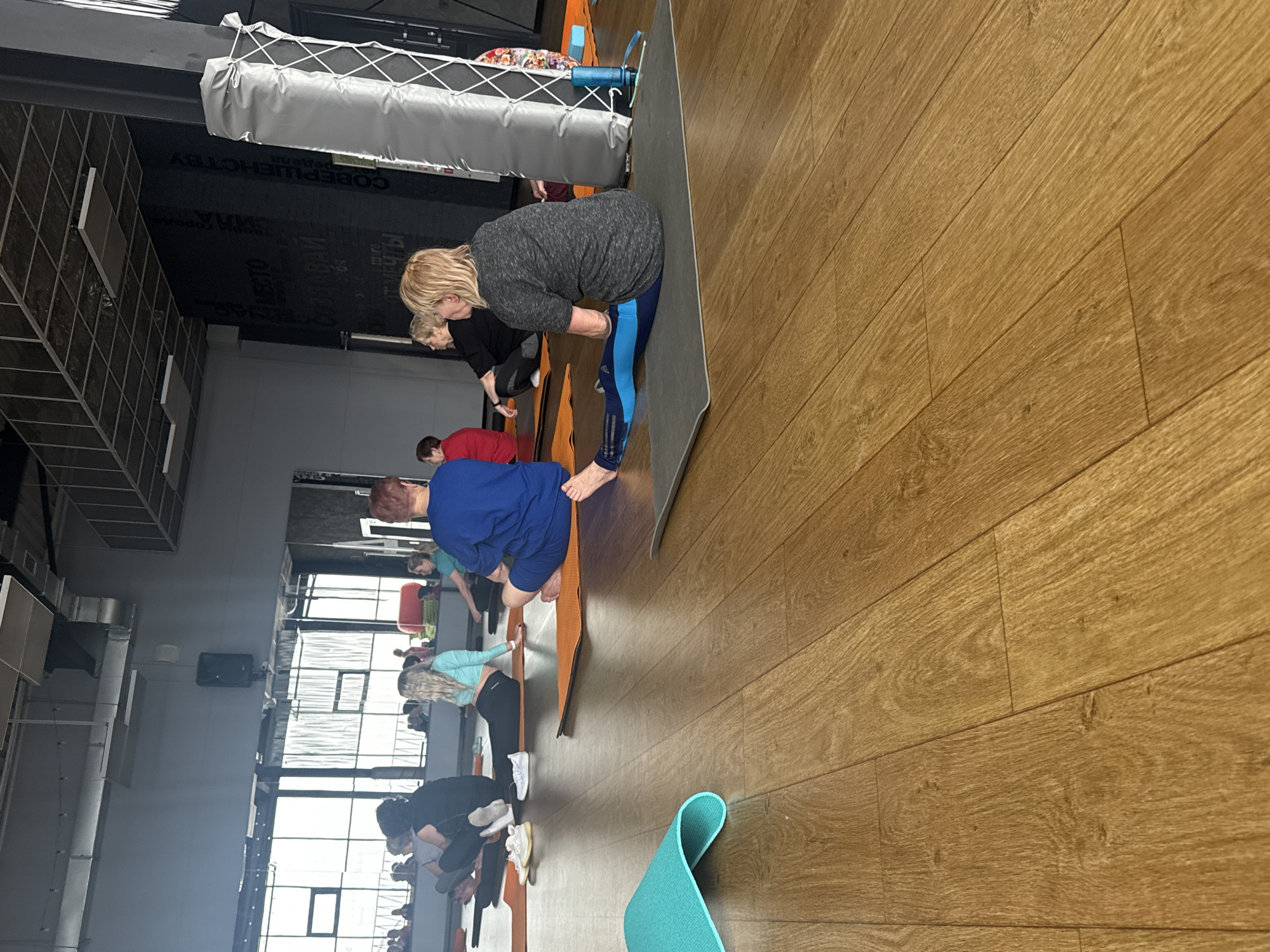 The image shows a yoga class taking place indoors, likely a fitness studio. There are at least 10 people visible, arranged in a row on yoga mats. They appear to be performing a forward bend pose. The studio has a wooden floor and large windows. A padded structure hangs from the ceiling. The location is Votkinsk, Russia, based on other contextual information.