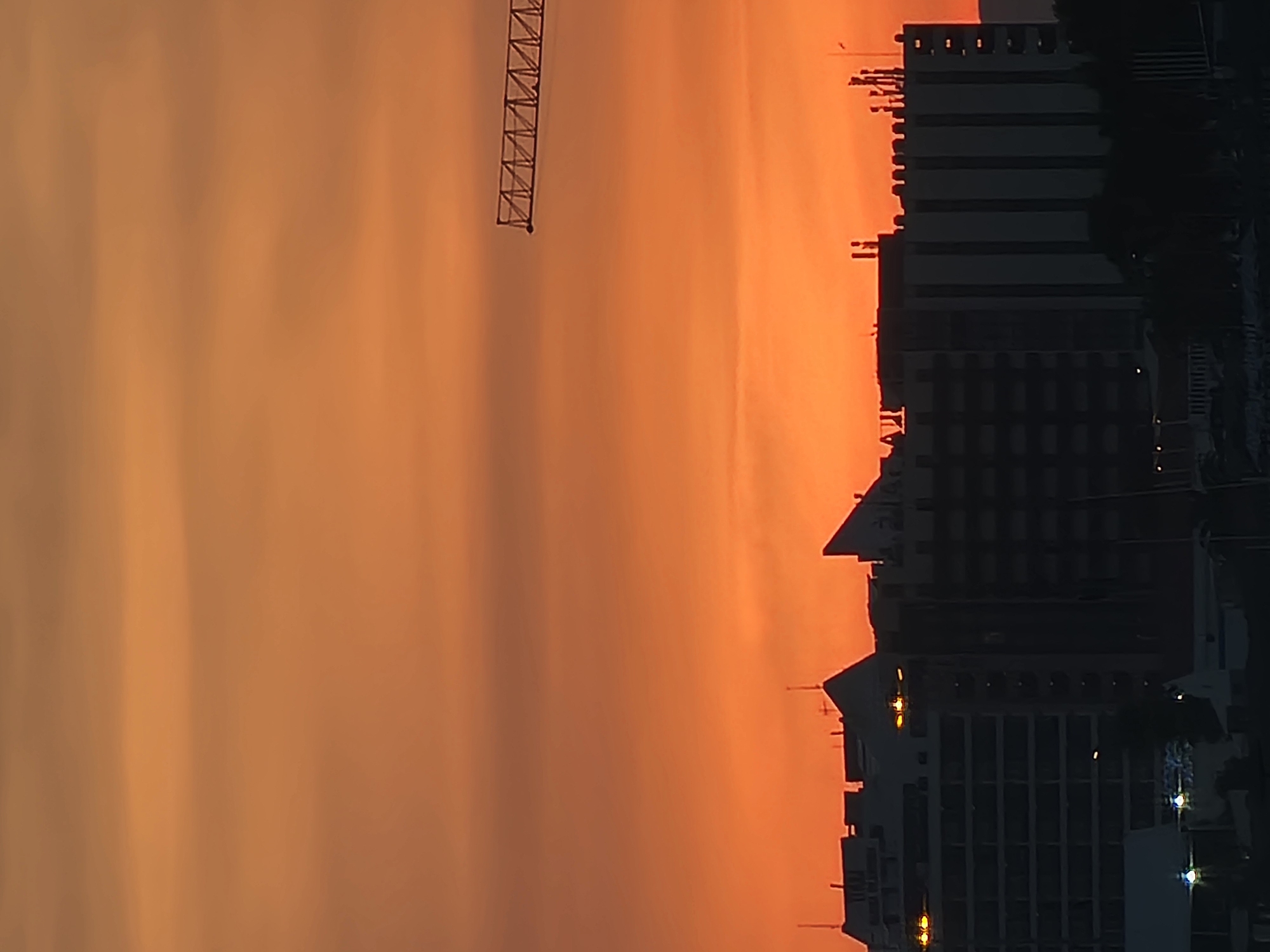 A silhouette of buildings and a crane is visible against a vibrant orange sky, indicative of a sunset in Quarteira, Portugal. The buildings are dark, some featuring distinctive architectural shapes and antennas on their rooftops. One building appears to have several windows lit from within. In the upper right portion of the image, a section of a construction crane's boom is visible, its lattice structure stark against the colorful sky. The sky itself is a gradient of orange and yellow, with some faint cloud formations. The scene is devoid of any visible people or ongoing actions.