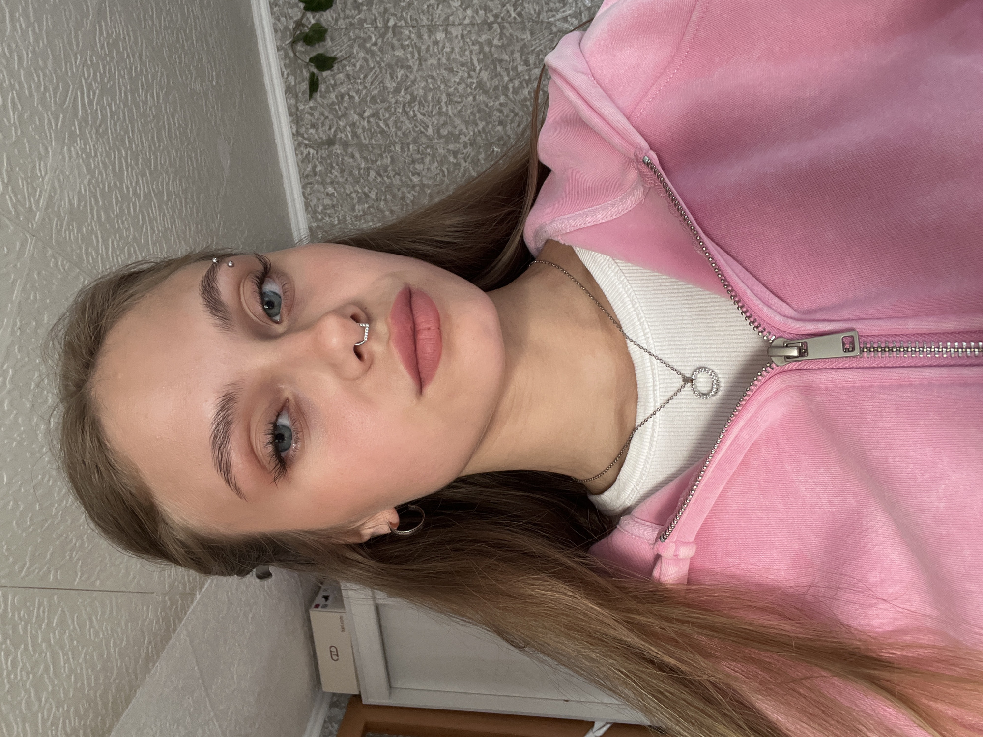 A young woman is lying down, facing upwards and angled slightly to the right. She has fair skin, light brown hair, and blue eyes. A metal piercing is visible in her right eyebrow, and she has a nose ring in her left nostril. She is wearing a pink zippered hooded sweatshirt over a white top. A silver necklace with a circular pendant is around her neck. The background is a textured wall, and part of a plant can be seen in the upper left corner. A small white cabinet is in the lower left corner. The location is unspecified, and there is no information in the image to indicate that the location is in the city of Фоки, Russia.