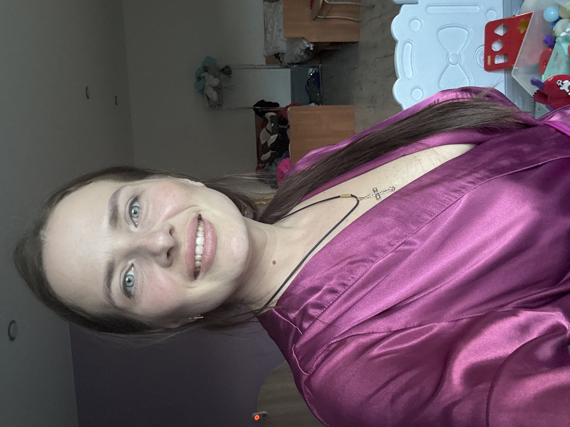 The image shows a close-up, vertically oriented, selfie-style shot of a young woman with light skin, blue eyes, and long dark brown hair. She is wearing a shimmering, deep purple robe. Around her neck, she has a thin black cord with a silver cross pendant. She is smiling broadly, her teeth visible. Her makeup includes blush on her cheeks. The background is slightly out of focus but suggests an indoor setting with some furniture, possibly a bedroom or living area, with items on shelves and a white plastic play structure. The lighting appears to be from artificial sources, creating a soft glow. The overall mood is cheerful and relaxed. There is no visible text in the image.