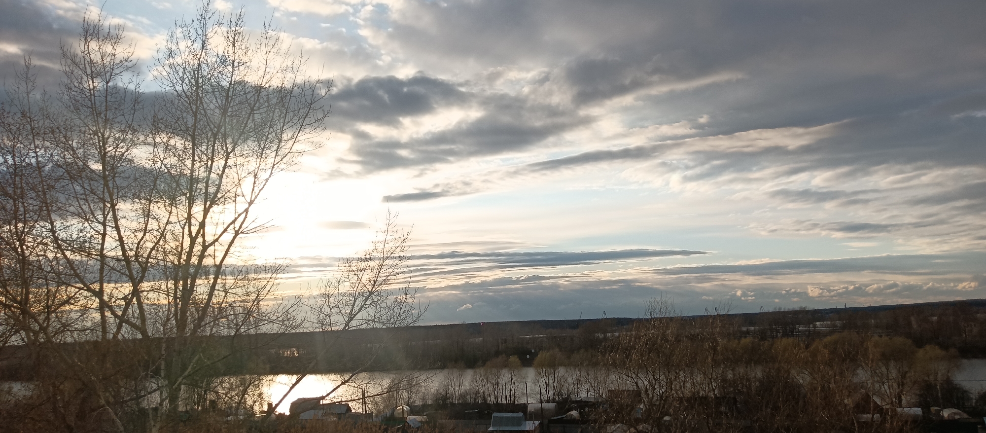 The image presents a serene landscape captured likely during either sunrise or sunset, characterized by a low, bright sun that illuminates the scene from the left. In the foreground, a dense arrangement of bare, leafless tree branches, typical of late autumn, winter, or early spring, dominates the left side of the frame, creating a striking silhouette against the luminous sky.
Beyond these foreground trees, a calm body of water, possibly a river or lake, stretches across the mid-ground. The intense sunlight reflects brilliantly off its surface, creating a golden shimmer. Along the distant banks of the water, more bare trees and scattered, low-lying structures, resembling small houses or sheds, are visible, suggesting a rural or semi-rural settlement.
The horizon in the background features a dark, undulating treeline, with faint hints of distant industrial elements like cranes or towers. The sky above is a prominent feature, filled with a dynamic array of clouds. These range from soft, bright whites and light blues near the sun to darker, more dramatic greys and purplish tones across the upper parts of the frame. The clouds appear layered, contributing to the atmospheric depth of the scene. The overall mood conveyed is one of peaceful tranquility, with the interplay of light and shadow enhancing the natural beauty. There is no visible text in the image.
