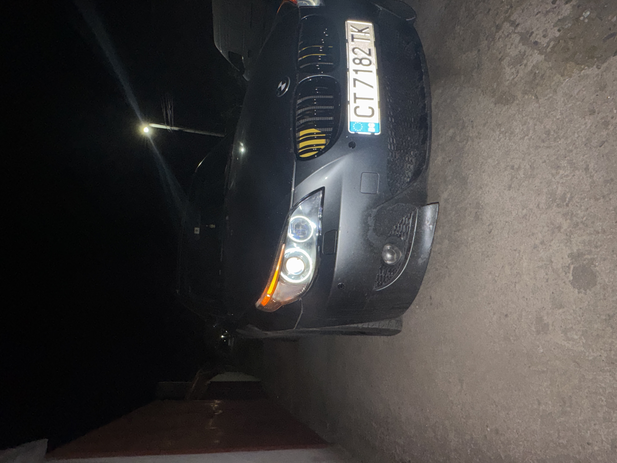 The image depicts the front of a dark-colored BMW, captured at night. The car's license plate reads "CT 7182 TK" and includes a Bulgarian flag detail, suggesting it is a Bulgarian vehicle. The vehicle has after market modification with yellow paint in between the grille bars. The location is Sarnevo, Bulgaria. The surrounding environment appears to be dimly lit, likely a street, with the ground covered in pavement.
