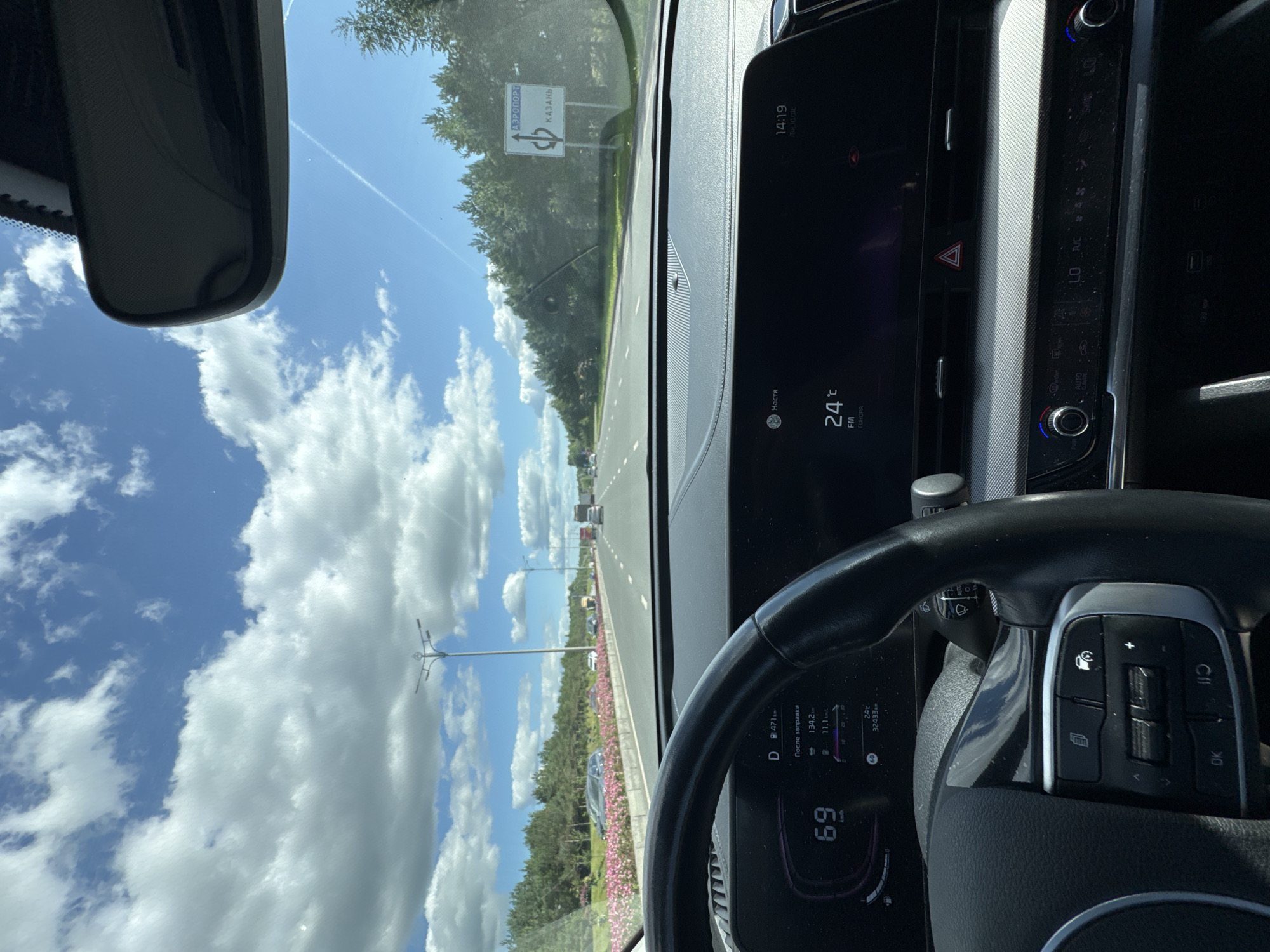 The image shows the interior of a car driving on a highway. The dashboard displays the current speed as 69 km/h, the outside temperature as 24°C, and the time as 14:19. A road sign on the right side of the highway indicates directions to "АЭРОПОРТ" (Airport) and "КАЗАНЬ" (Kazan). The highway is lined with trees and flowers, and the sky is blue with white clouds. There are other vehicles visible on the highway in the distance. The steering wheel and part of the car's interior are in the foreground. The location is the M7 highway approaching Kazan, Russia.