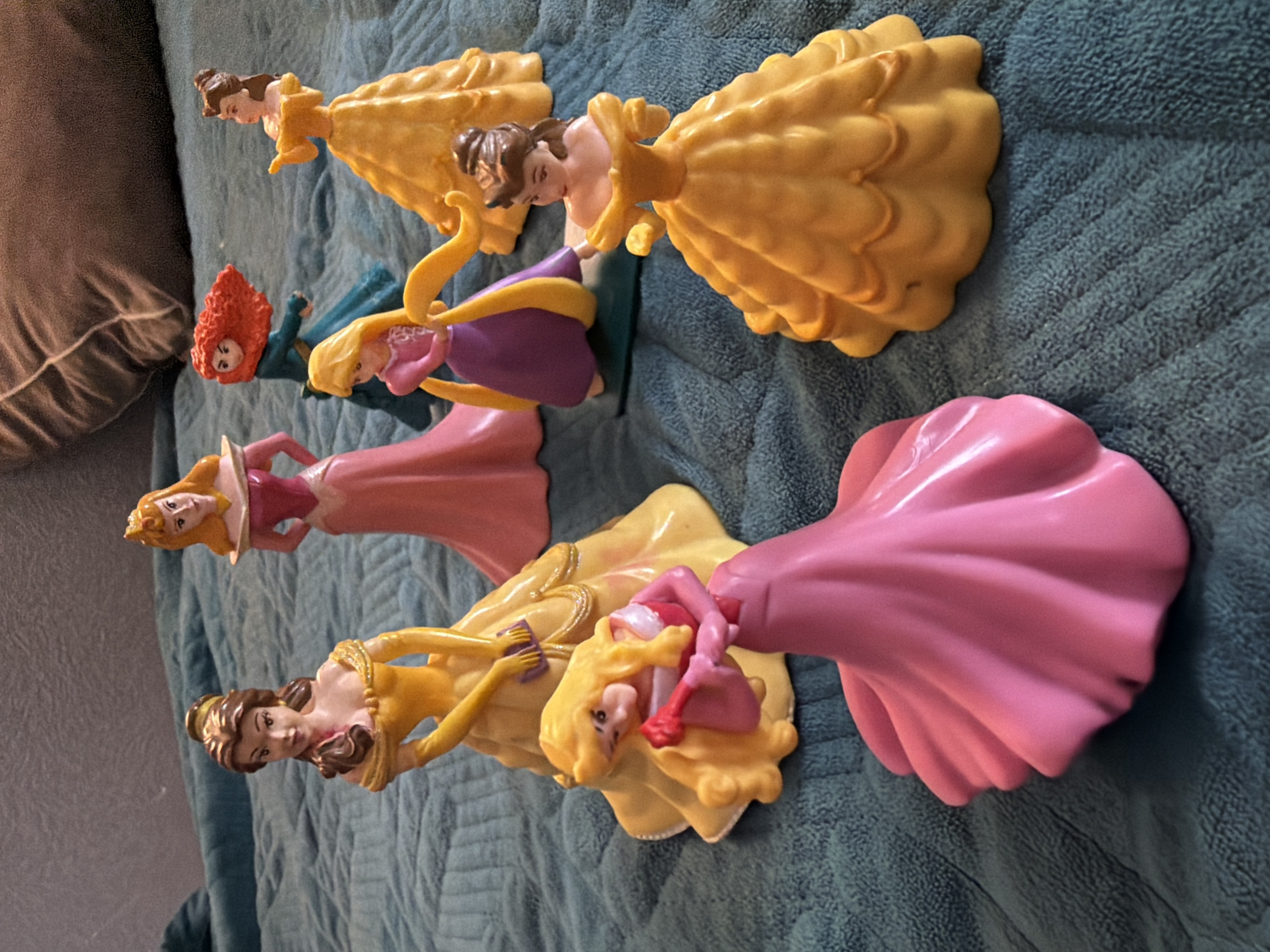 The image shows six Disney princess figurines arranged on a blue textured blanket.
From top to bottom, the princesses appear to be:
1. Belle in her yellow ballgown
2. Rapunzel in her purple dress, holding a paint palette
3. Merida in her teal dress
4. Aurora (Sleeping Beauty) in her pink dress
5. Belle in her yellow dress, holding a paint palette and cleaning
6. Snow White in her dress
The princesses are arranged in varying orientations on the blanket.
There is a pillow in the upper left corner of the image.
The location is not evident.