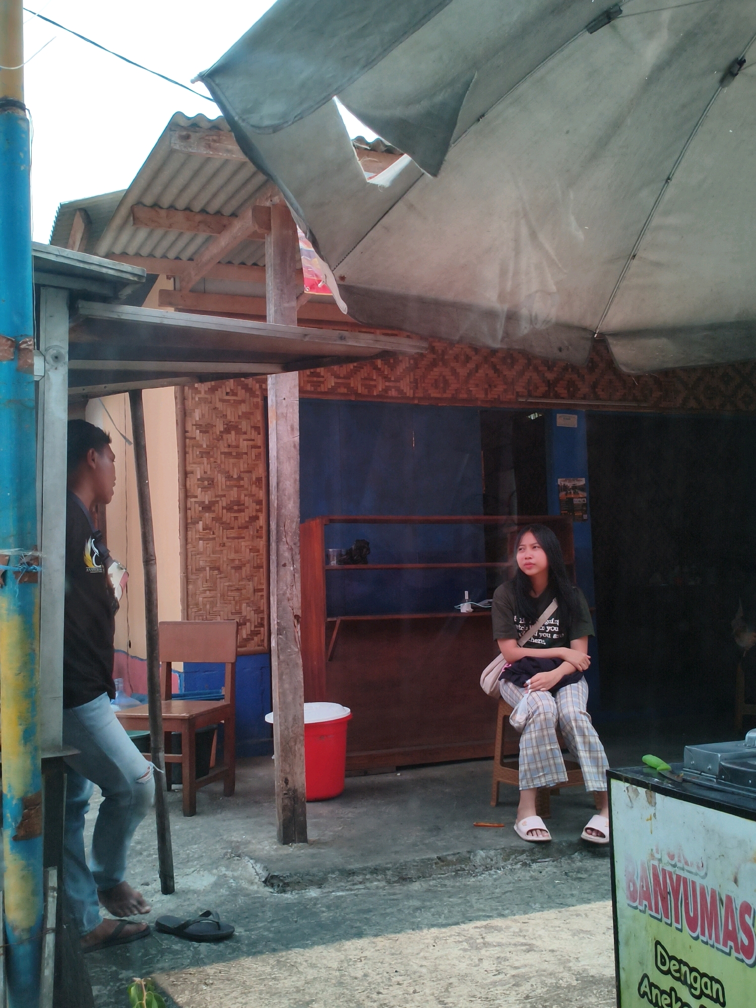 A young man and a young woman are standing and sitting, respectively, in an outdoor marketplace stall in Sukaraja, Indonesia. The stall has a corrugated metal roof with a torn, light-colored tarp stretched over it. The walls appear to be made of bamboo weaving and dark blue tarpaulin.
The man, wearing a black t-shirt and ripped blue jeans, stands near a blue and yellow pole on the left, facing away from the camera. He is barefoot on one foot, and wearing a sandal on the other. A wooden chair is behind him.
The woman, wearing a dark t-shirt with white text and plaid pants, sits on a wooden stool on the right. She has a light-colored crossbody bag over her shoulder and her arms are crossed. She is wearing white slides.
In the background, there are shelves with some items on them and a red trash can. A food display case with text written on it is visible in the foreground on the right. The ground is a concrete surface. The sky is visible in the upper part of the image.