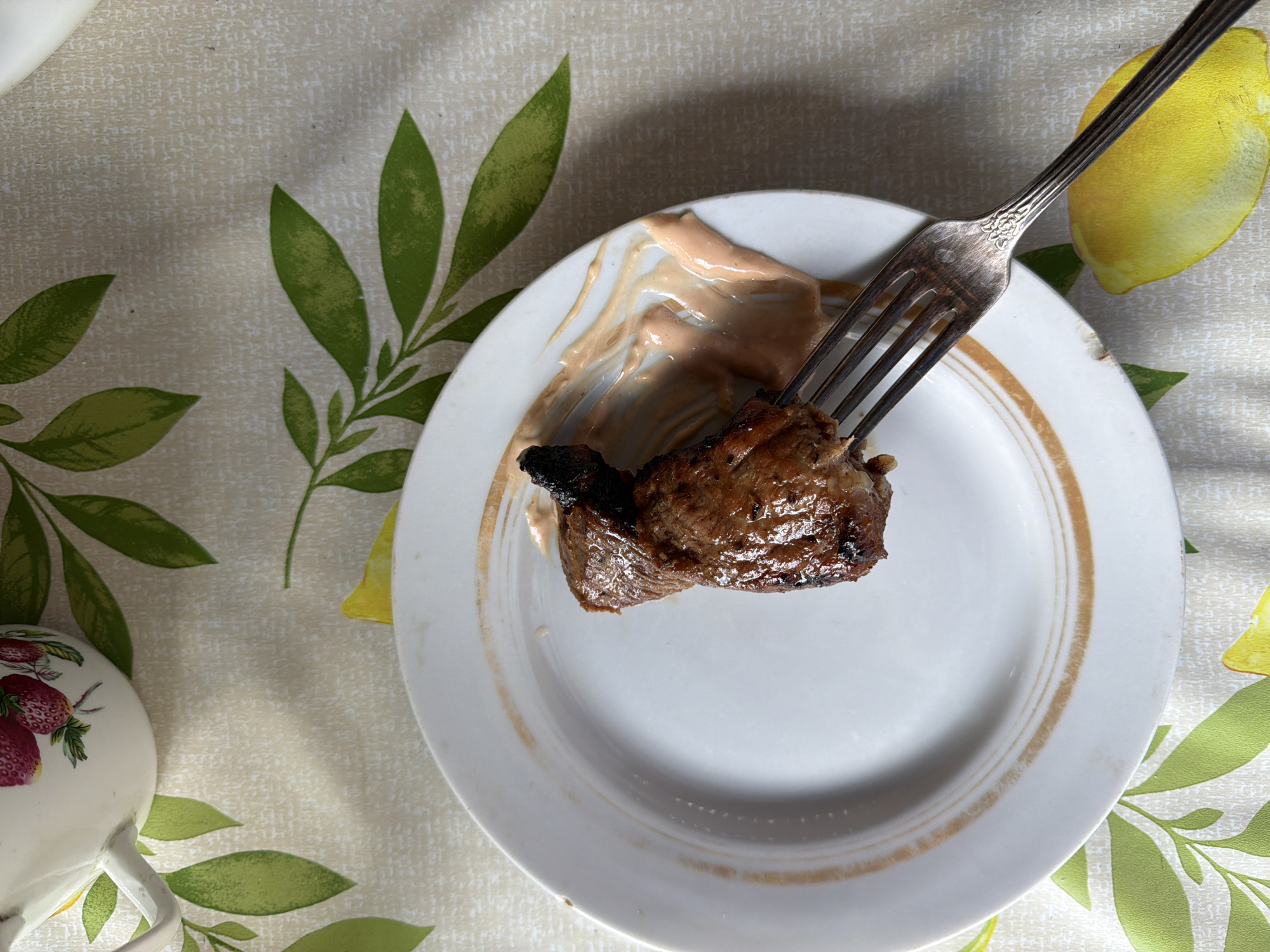 A close-up, overhead shot shows a single piece of grilled meat impaled on a fork, resting on a white plate with a brown decorative rim. The meat is dark brown with some charring. A swirl of pinkish sauce is smeared on the plate next to the meat. The plate is situated on a light-colored tablecloth patterned with green leaves and yellow lemon slices. In the upper left corner, a portion of a white teapot with a strawberry design is visible. The image does not contain any specific information about the city of Kurgan, Russia.