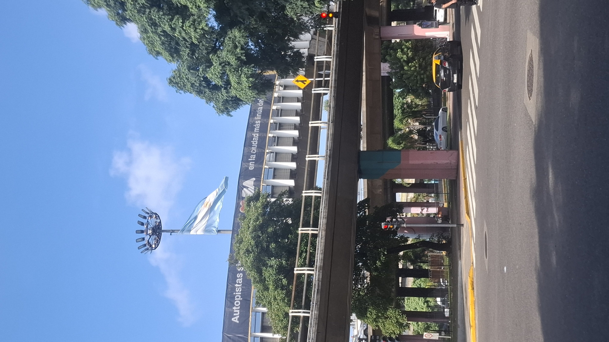 The image depicts a street scene in Buenos Aires, Argentina. An elevated highway structure is visible above the street, with a road running parallel to it. A banner on a building reads "Autopistas en la ciudad más linda del mundo." The Argentine flag is flying on a pole. A yellow taxi and other cars are on the road. Pedestrians are present. A traffic light is showing red. The sky is blue with some clouds, suggesting daytime. Trees are also visible, contributing to the urban landscape.