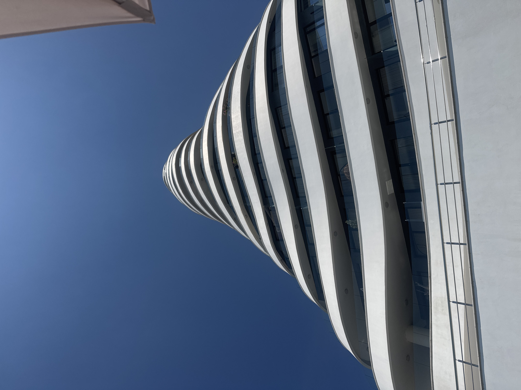 The image shows a low-angle, wide shot of a modern, curved building against a clear blue sky. The building features a distinctive design with undulating white facade elements that create a wave-like pattern. These elements curve and twist, giving the structure a dynamic and almost sculptural appearance. Large glass windows are visible between the white architectural features, reflecting the sky and appearing dark in contrast. The perspective emphasizes the height and sweeping lines of the building.
In the upper left corner of the frame, a portion of a light-colored, rectangular object, possibly an umbrella or awning, is visible, casting a shadow and suggesting an outdoor setting. The lighting indicates it is daytime, likely with bright sunlight given the clear sky and the strong contrast between light and shadow on the building. No people are clearly visible, and the focus is entirely on the architecture and the sky.
The building's sleek, contemporary design and the bright, unobstructed sky suggest a location with modern infrastructure and a clear, pleasant weather day. The presence of the partial umbrella hints at a public space or an area designed for outdoor enjoyment. The overall impression is one of modern design and a serene, bright atmosphere. The image's orientation is vertical, with the building curving upwards from the bottom left towards the top right. No text is visible in the image.