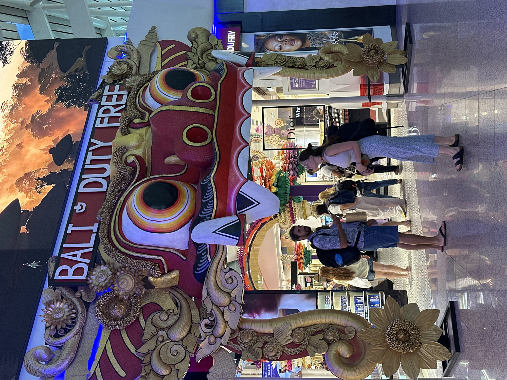 The image shows a scene at Bali's Ngurah Rai International Airport in Kuta, Indonesia. The foreground features a large, ornate display with the words "BALI DUTY FREE" prominently displayed alongside a depiction of a traditional Balinese mask with golden accents. Behind this display are people, seemingly tourists, walking in different directions. Some appear to be looking at shop windows, including brands like Dior and Estée Lauder, which are visible in the background. The floor has a polished, reflective surface, suggesting a modern airport setting. The lighting is bright, likely from overhead fixtures and illuminated shop displays.