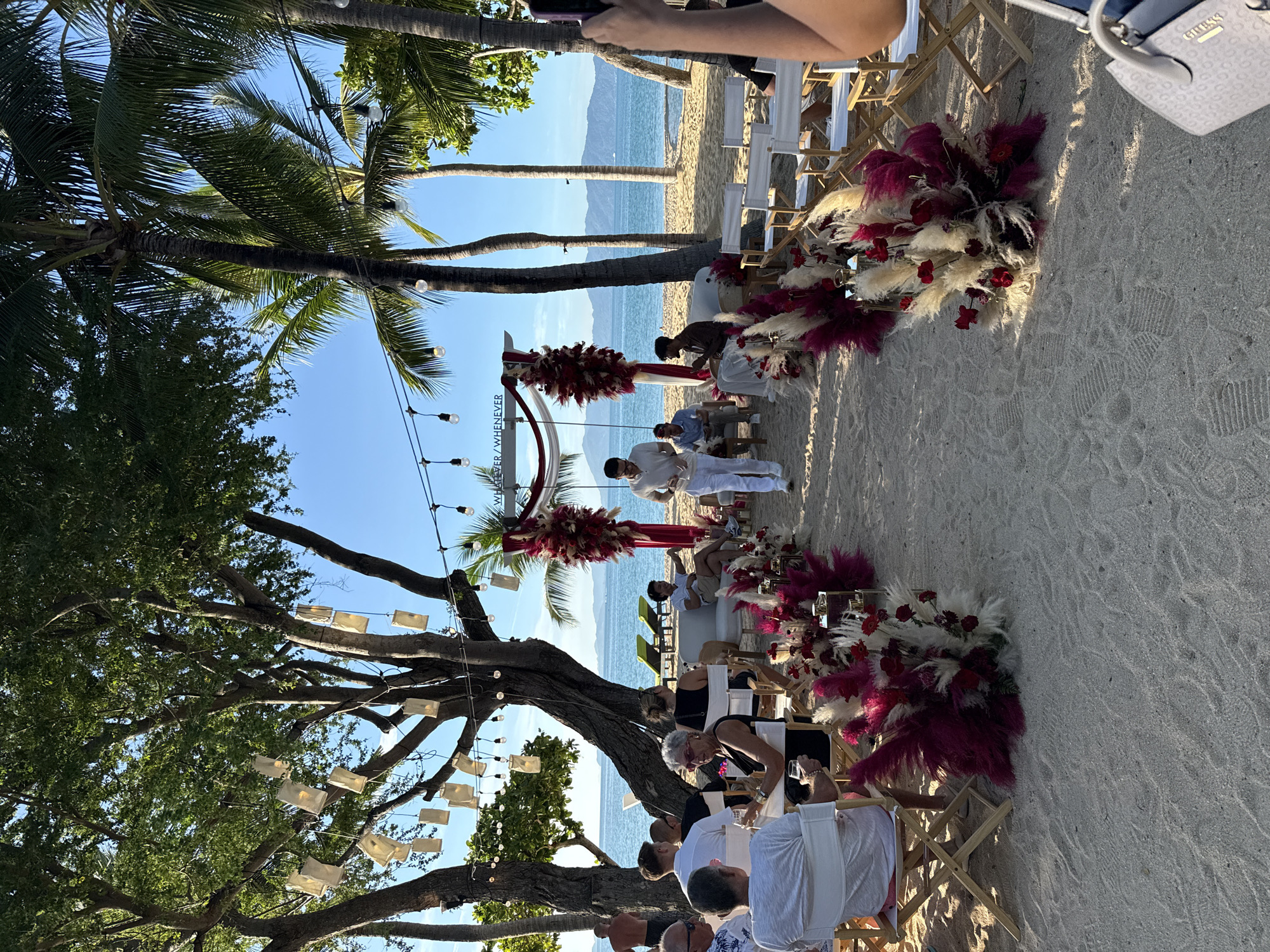 This is a medium shot, outdoor photograph taken on a sandy beach in Baan Bo Phut, Thailand. The scene depicts a wedding ceremony setup with a decorated archway and floral arrangements along an aisle. Several people are present, seated on chairs and lounges, facing towards the archway. The ocean and distant islands are visible in the background, under a clear blue sky. The lighting suggests it is late afternoon, with the sun casting long shadows.