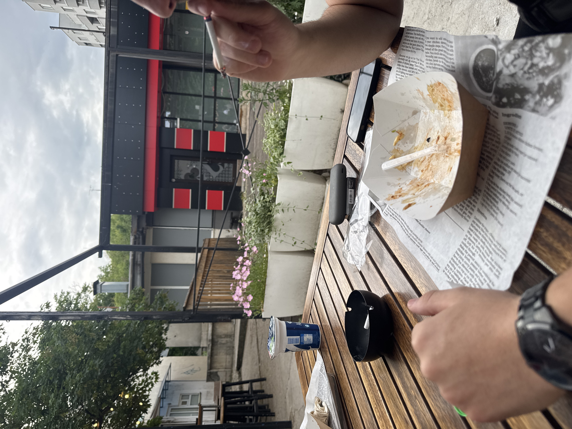 The image shows a view from a wooden table at an outdoor cafe. A person is holding a lit cigarette, and another person is seated at the table with a watch on their wrist. A takeout box with leftover food is on a newspaper, along with a yogurt cup and a black container. The cafe appears to be in an urban area, likely in Ruse, Bulgaria, based on context. The backdrop includes a building with a black and red facade and surrounding greenery. Other tables and chairs are visible in the background.