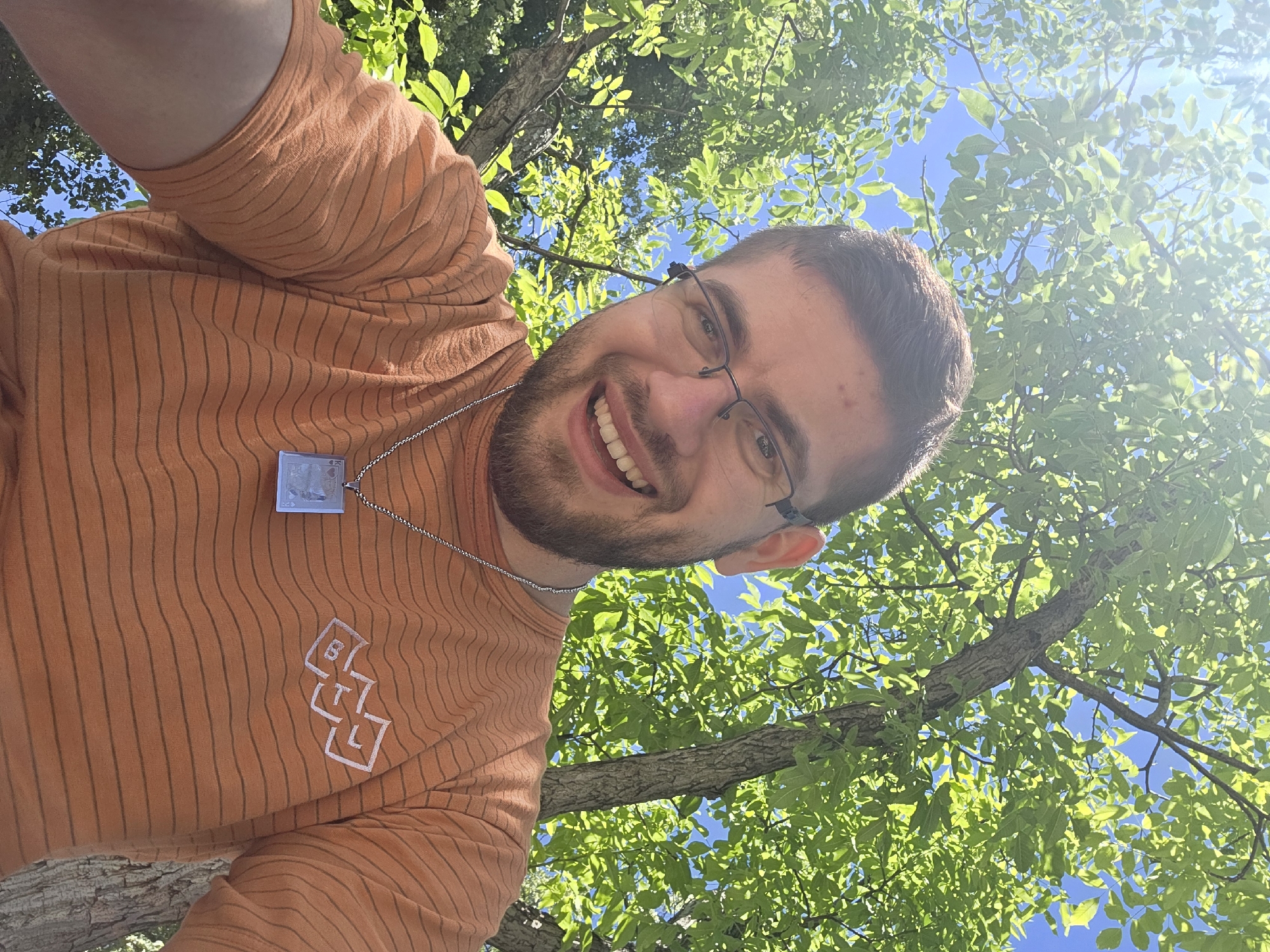 A man is looking down at the camera, smiling, with a tree overhead. He is wearing an orange, striped shirt with a logo on it, and a chain necklace with a rectangular pendant. He has glasses and a beard. The background consists of green leaves and a blue sky. The location cannot be determined from the image.