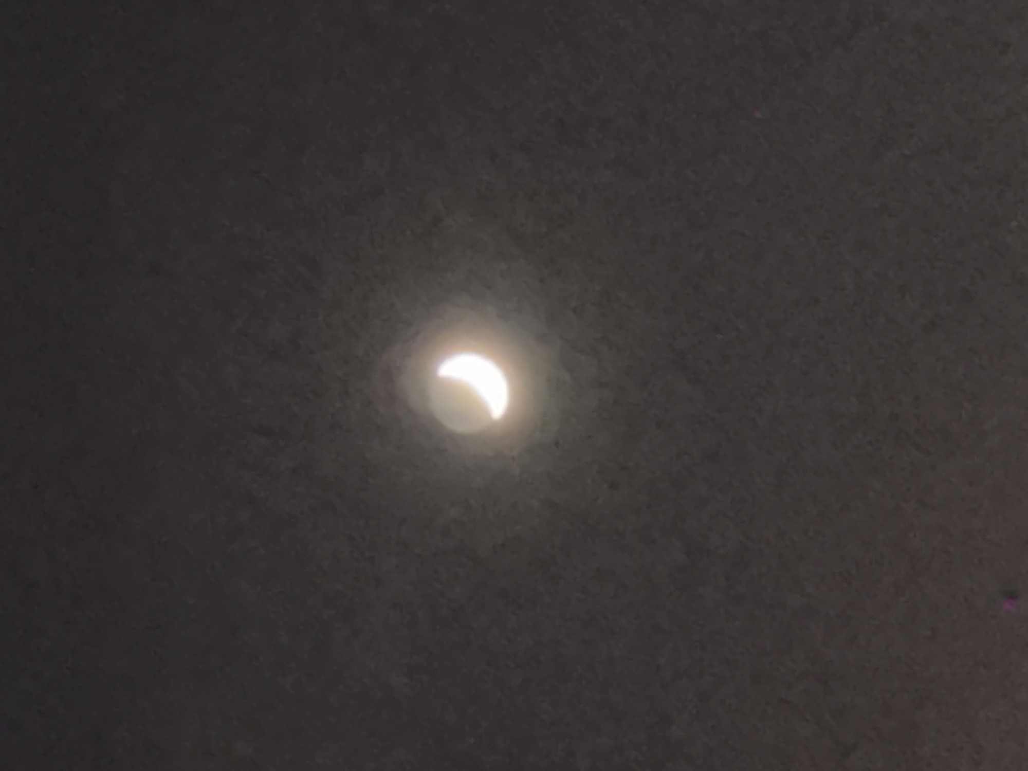 The image displays a partial eclipse, characterized by a bright, luminous crescent-shaped object centrally positioned against a dark, textured background. The crescent is intensely bright and appears white with a subtle glow, suggesting it is a view of the sun during a partial solar eclipse, likely observed through a protective filter or projected onto a surface. The dark background is uniform, showing a faint grainy or cloudy texture, typical of either a filtered sky or the surface used for projection. No other celestial bodies, terrestrial objects, or distinct features are visible in the frame. The scene focuses exclusively on the astronomical event. There are no people, discernible emotions, or visible text within the image. The time of day is indicated as daytime during the eclipse event, with clear conditions allowing for this observation. The provided location context is Khimki, Russia.