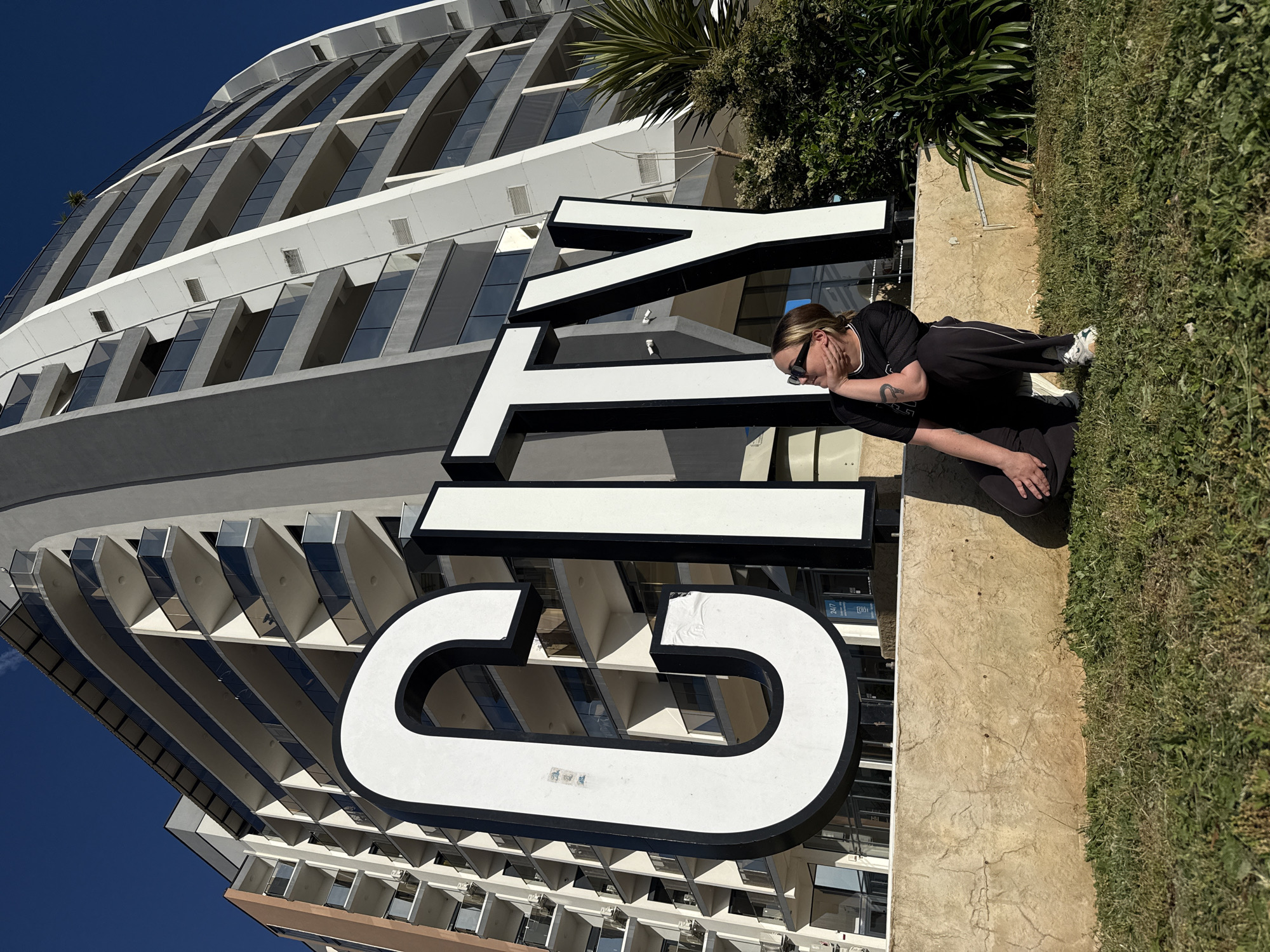 A wide, rotated, outdoor shot features a person in dark clothing kneeling on tan concrete next to a "CITY" sign in large white letters with black outlines in front of a tall building. The person is fair-skinned, wearing sunglasses, with their left hand covering part of their face and a visible tattoo on their left arm. The building is multi-story with a modern architectural design, featuring many windows and white/grey panels. A small portion of green foliage is visible near the top of the frame, and a blue sky is in the background. The location is Bar, Montenegro.