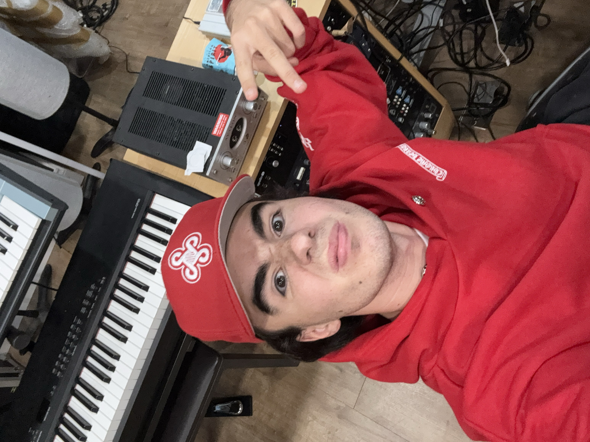 In an indoor setting, a young Caucasian male with dark hair, wearing a red baseball cap and a red hooded sweatshirt, is taking a selfie. He is making a playful or surprised facial expression, with his eyebrows raised and mouth slightly open. His right hand is raised, with his index and middle fingers extended in a peace sign gesture. The background reveals a music studio environment. Two black and white electronic keyboards are visible, one stacked above the other. A wooden desk holds various audio equipment, including a silver rackmount device with knobs and buttons, a black piece of equipment with ventilation slots, and several coiled cables. The floor appears to be light-colored wood or laminate. The overall scene suggests a casual and creative atmosphere.