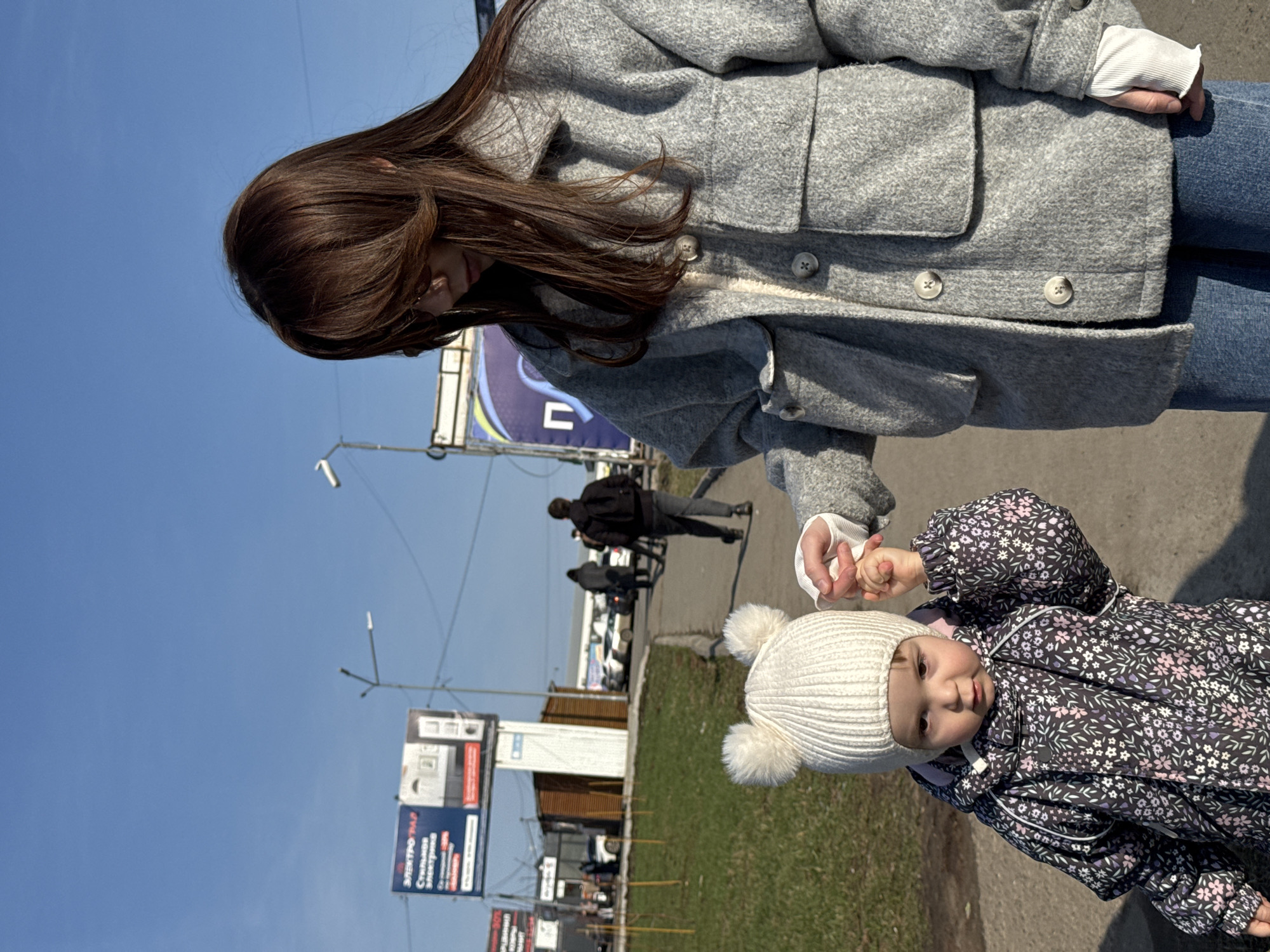 A woman and a child are shown in an outdoor setting, likely a street in Chaikovsky, Russia. The woman, partially visible from the chest up, has long, brown hair and is wearing a gray coat and blue jeans. She is holding the hand of a toddler dressed in a dark, floral-patterned snowsuit and a white knit hat with pom-poms. In the background, there are streetlights, billboards with Russian text, and figures walking on the sidewalk. Cars are parked in the distance. The sky is clear and blue.