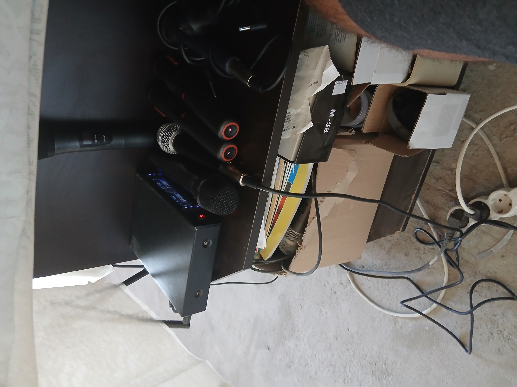 A top-down, slightly rotated shot reveals a cluttered shelf in Vetren Dol, Bulgaria. Visible on the left are two black wireless microphones and a black receiver with visible interface lights. To the right of the receiver, more microphones lie among boxes and cords, including a box labeled "M-58" and some Cyrillic text. The shelf is overflowing with items, and the floor beneath is littered with electrical cords and a power strip. A portion of a person's arm, clad in gray fabric, appears in the top right corner of the frame.