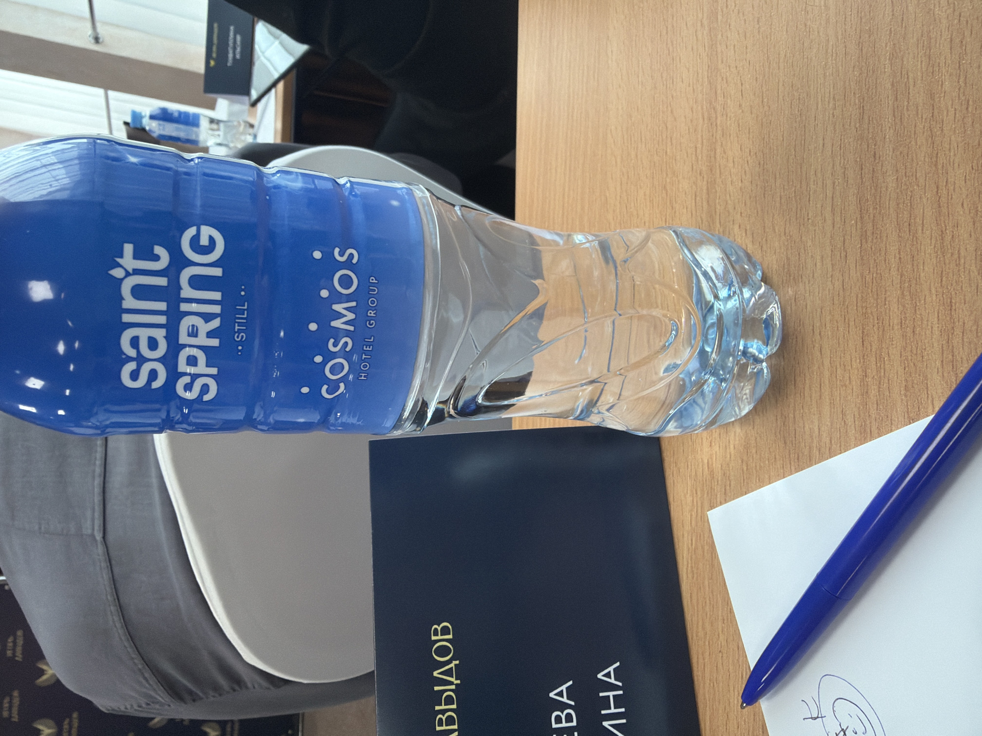 A slightly rotated, close-up shot depicts a table scene. A bottle of Saint Spring still water, branded with Cosmos Hotel Group, lies horizontally. A dark blue booklet with gold Cyrillic text sits partially under the water bottle. A white sheet of paper with a blue pen resting on it is present, with a faint sketch visible on the paper. The table surface appears to be a light-colored wood grain. The scene suggests a meeting or conference setting. Without additional information, determining the exact location in Pervomaysky, Russia is impossible.