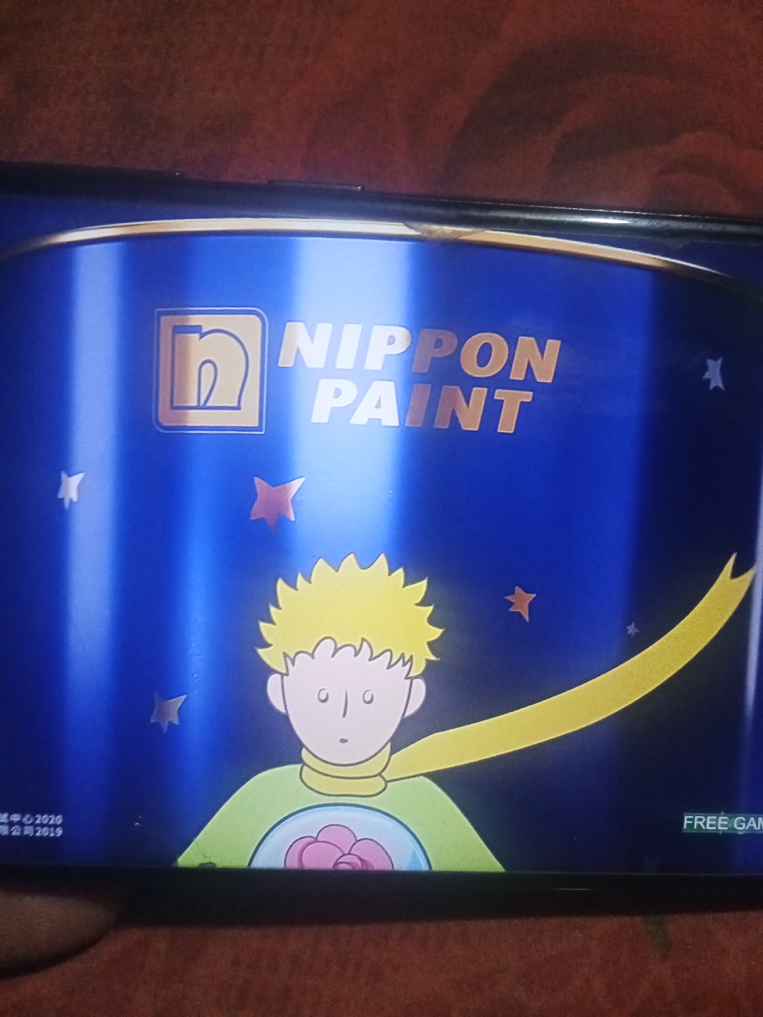 The image shows a screen, likely from a mobile device or television, displaying a graphic. The graphic features a blue background with several stars. In the upper portion of the screen, the text "NIPPON PAINT" is prominently displayed in large, stylized letters. To the left of the text is a logo consisting of a square with rounded corners containing the letter "n" in a lighter shade. Below the text and logo, a cartoon character resembling the Little Prince is depicted. The character has spiky yellow hair, wide eyes, and is wearing a green shirt and a yellow scarf. A large yellow curved shape, possibly representing a comet or a scarf, extends from the character across the screen. In the bottom left corner of the screen, some text is visible, appearing to be copyright information, with dates "2020" and "2019". In the bottom right corner, the words "FREE GAME" are displayed. The screen itself is dark-edged and rests on a textured surface that appears to be brown fabric. The lighting suggests the screen is illuminated. The provided image is an indoor shot of the screen, with no discernible elements of Buketglumpang, Indonesia.