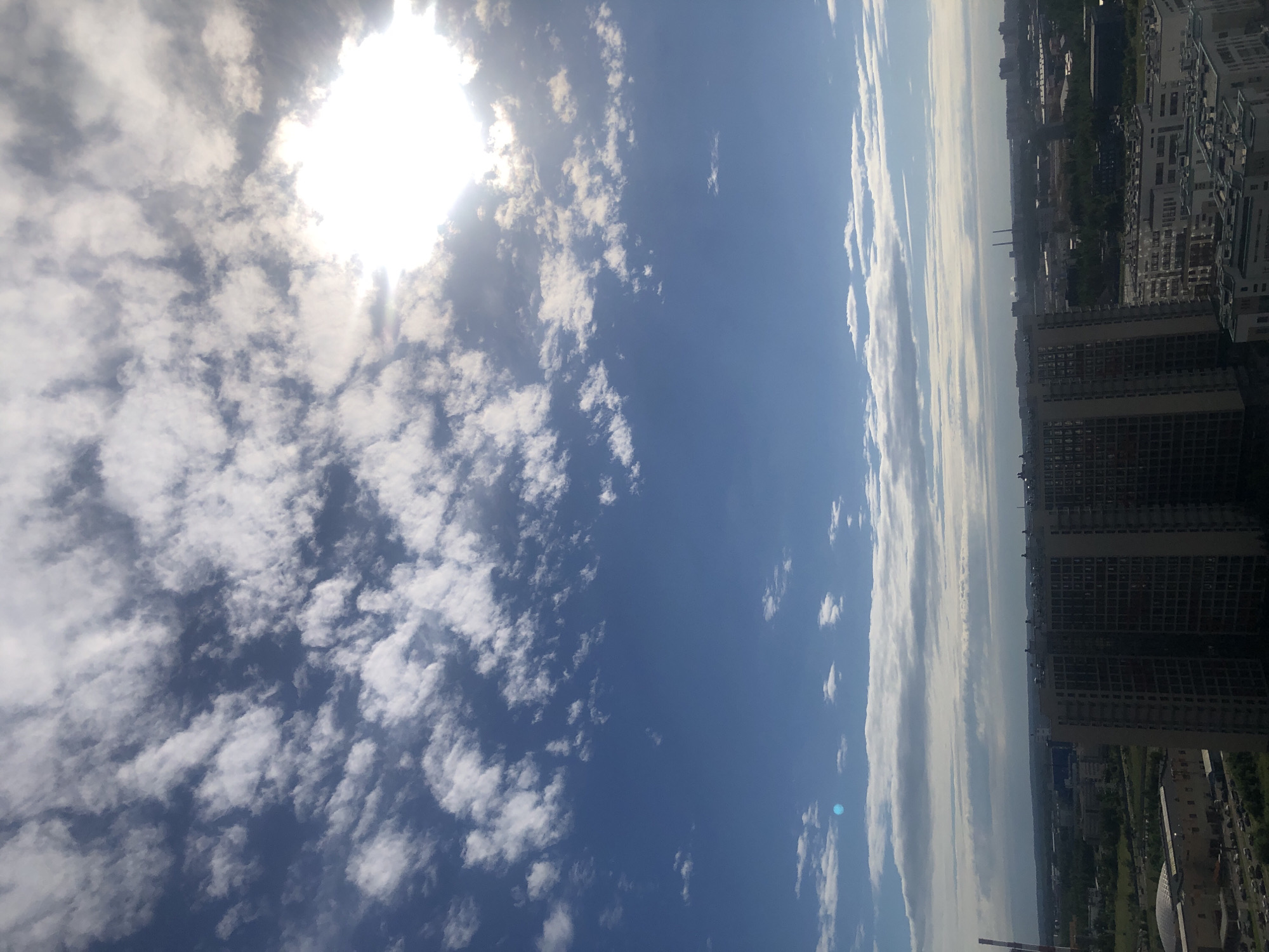 This is a vertical, low-angle shot of a cityscape under a cloudy blue sky. The sun is visible in the upper left corner, partially obscured by white, fluffy clouds, suggesting it's daytime. The sky takes up the majority of the frame, with scattered clouds creating a dynamic, textured ceiling.
On the right side of the image, a dense urban environment unfolds. Tall, modern apartment buildings with numerous windows dominate the foreground and midground. These buildings are dark and appear to be made of concrete or a similar material. Behind them, a sprawling cityscape with more buildings, green spaces, and what appear to be industrial structures stretches towards the horizon. The overall impression is of a busy, developed urban area.
The lighting suggests it's not the peak of midday, as the sun's glare is intense but not overwhelmingly bright, and some areas of the cityscape are in shadow. The weather appears partly cloudy, with a mix of bright sunlight and cloud cover. There are no people or specific objects that stand out besides the buildings. No text is visible in the image. The scene evokes a sense of scale and the vastness of an urban landscape beneath a wide, dynamic sky.