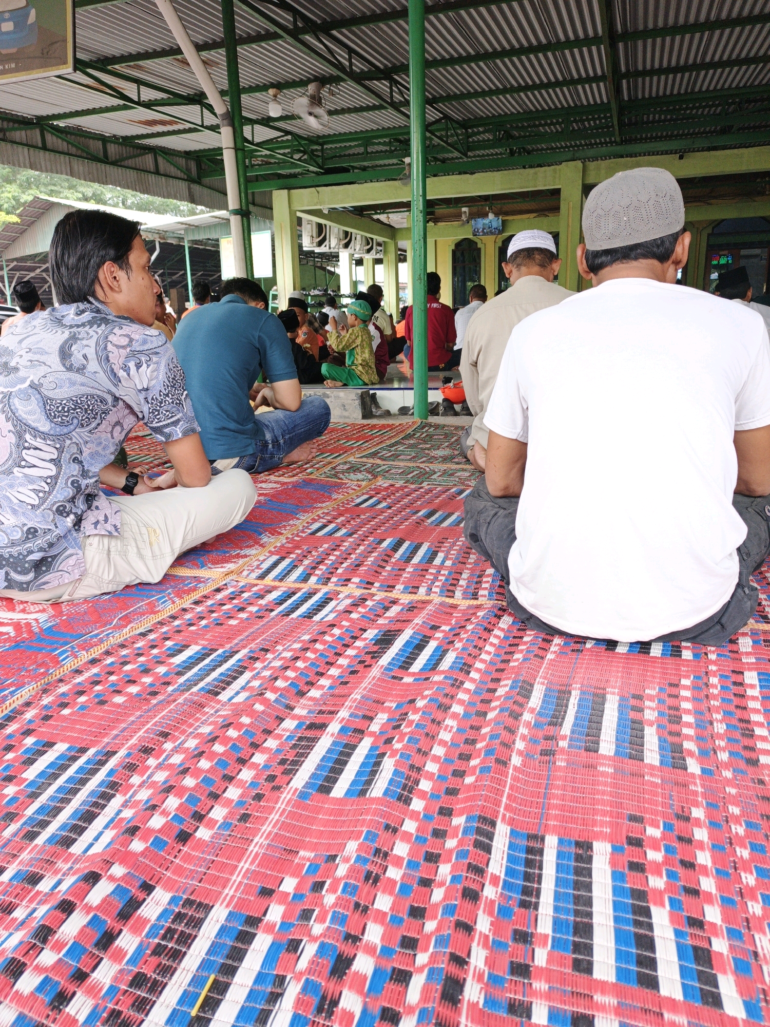 In the city of Manunggal, Indonesia, a group of men are seated on a red, white, and blue woven mat within an open-air structure. The structure's roof is made of corrugated metal supported by green metal beams and white poles. The men are facing away from the camera, engaged in prayer or contemplation. The man in the foreground on the right wears a white t-shirt and a grey skullcap. To his left, a man in a teal shirt and blue jeans is also seated. In the mid-ground, several other men are visible, some wearing traditional attire. The environment appears to be a place of worship or a community gathering space, indicated by the presence of religious symbols and the communal seating arrangement. The overall atmosphere is serene and orderly.