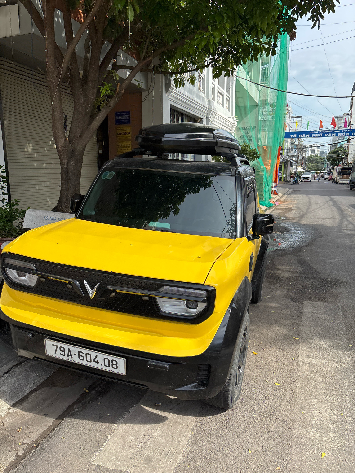 A yellow electric vehicle, possibly a VinFast VF 8, is parked on the side of a street in Nha Trang, Vietnam. The car has a black roof rack and black fender flares. Its license plate reads "79A-604.08". The street appears to be paved with asphalt and there is a sidewalk with a concrete bench. Buildings line the street, with one building under construction, covered in green netting. Several flags are visible in the distance, and other vehicles, including cars and trucks, are driving on the street. A tree with dense foliage stands to the left of the car. The sky is partly cloudy.