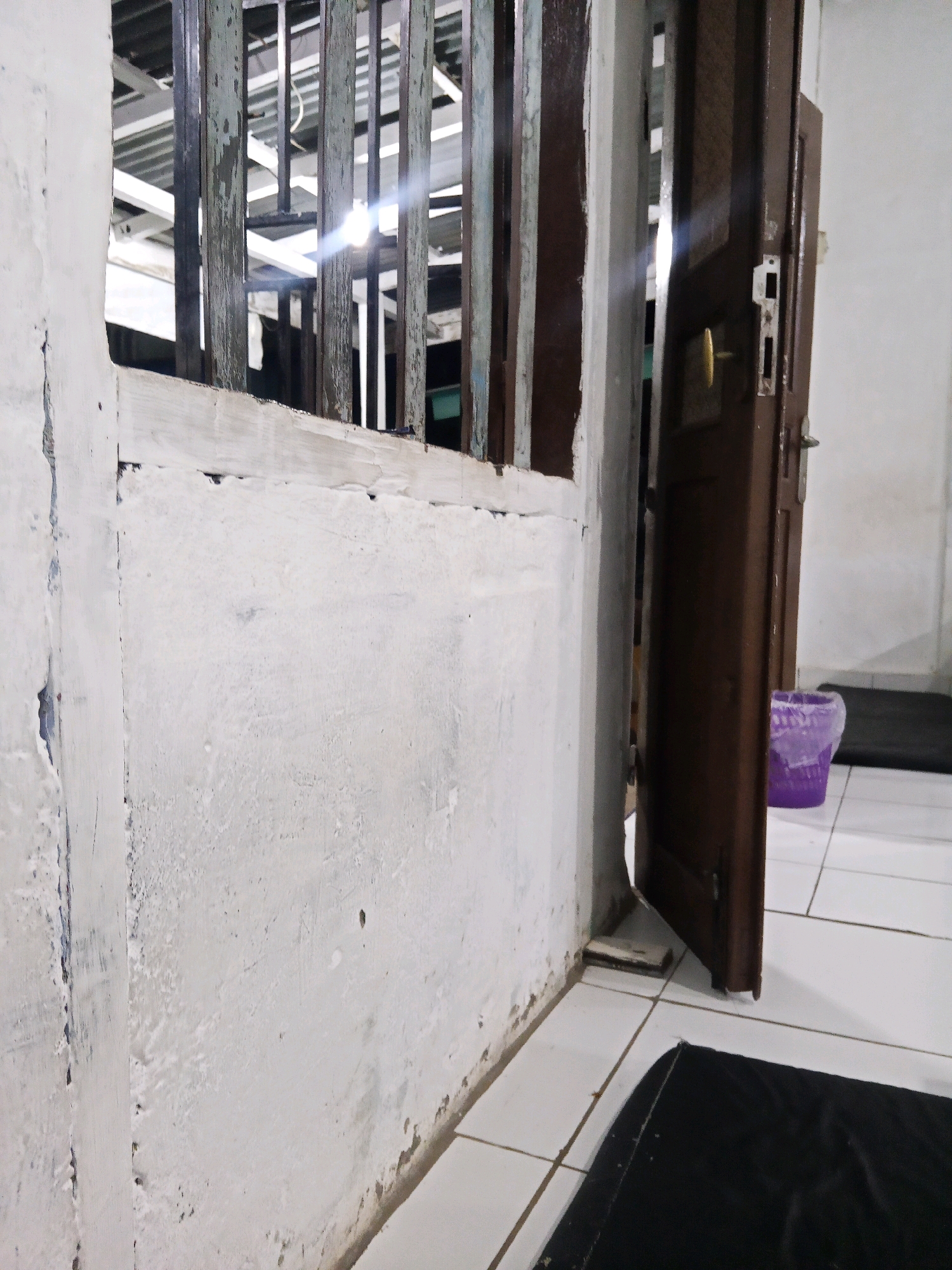 The image shows a close-up view of a doorway and a white concrete wall in Lubuklinggau, Indonesia. The doorway is made of dark brown wood and is slightly ajar, revealing a glimpse of what appears to be a room beyond. A metal grate, possibly part of a window or fence, is visible to the left of the doorway. The wall is textured and shows signs of age and wear, with some peeling paint and discoloration. The floor is tiled with white squares. A purple bucket and a black mat are also visible in the scene. The lighting suggests it is either dusk or dawn, or the area is indoors with artificial lighting.