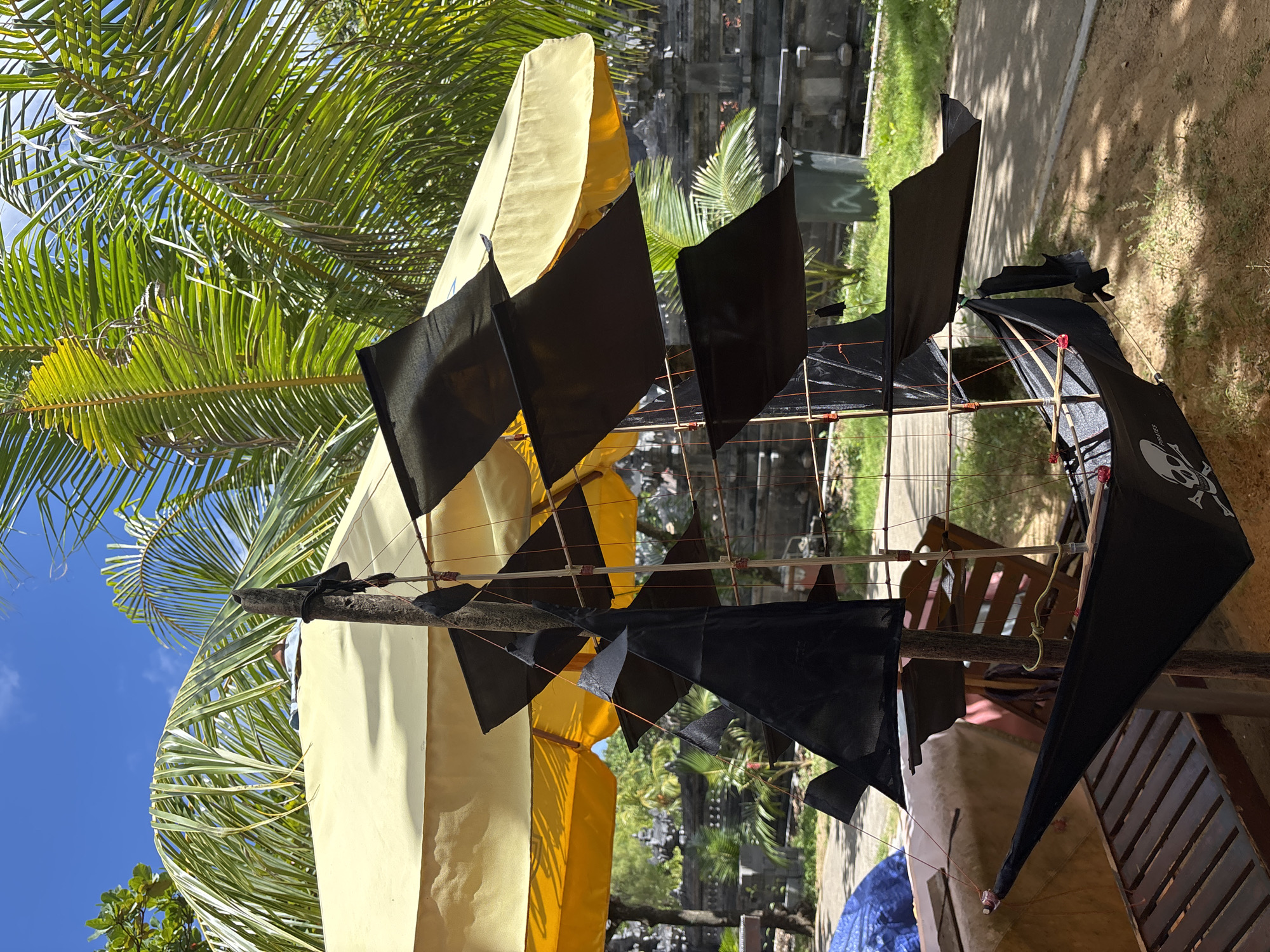 The image depicts a partially constructed model pirate ship outdoors in Kuta Selatan, Indonesia. The ship is made of bamboo sticks and fabric sails. The sails are a mix of yellow and black fabric, with one black sail featuring a skull and crossbones. The ship is resting on a wooden bench or table. In the background, there are palm trees, a traditional Balinese temple structure, and some vegetation. The sky is visible in the upper left corner, indicating a bright, sunny day.