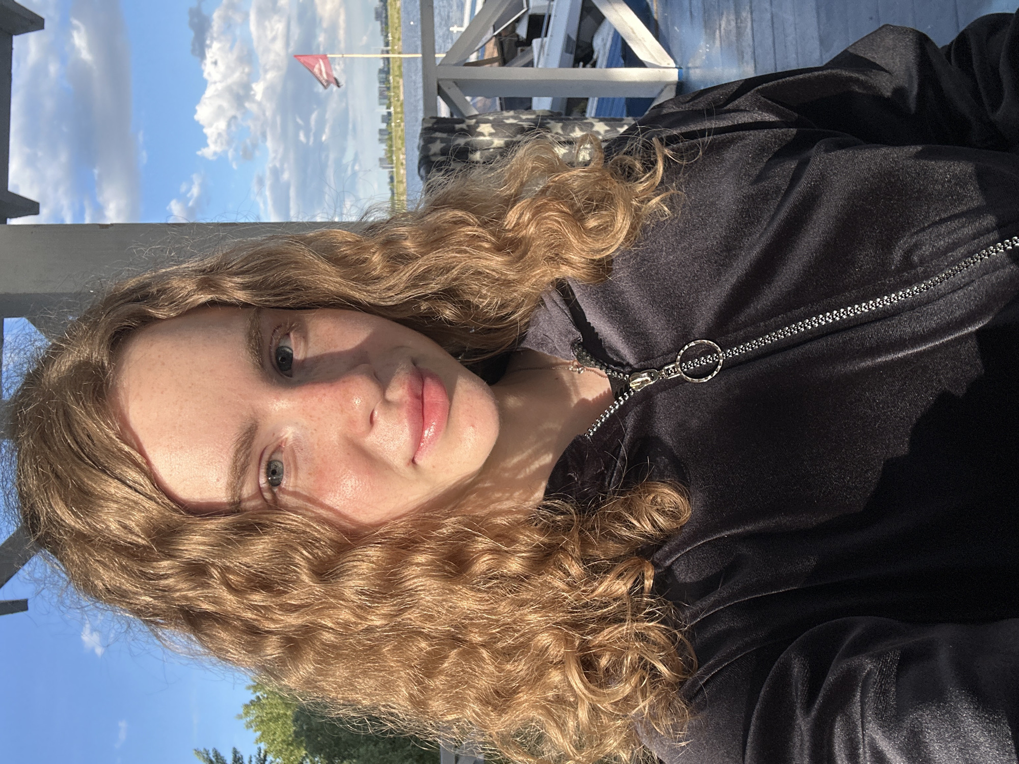 A young woman is posing for a selfie outside on a sunny day. She has curly blonde hair, fair skin, and is wearing a dark jacket with a zipper. In the background, there is a body of water, possibly a lake or river, with a red flag visible on a pole. The skyline of Kazan, Russia is visible in the distance. The woman is likely on a dock or pier, as indicated by the wooden structure and railings. The lighting suggests it is daytime.