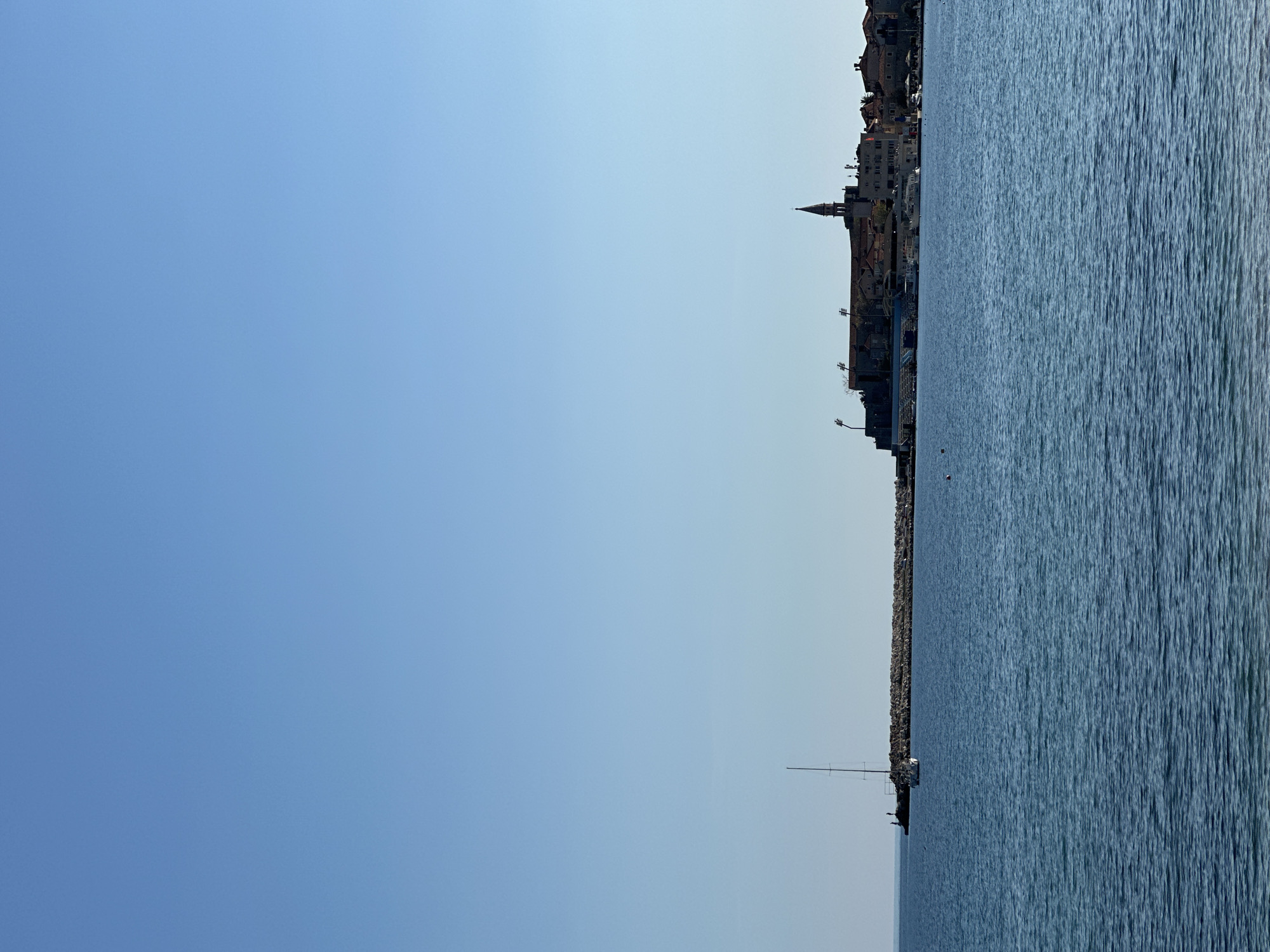 The image shows a view of the city of Budva, Montenegro, from the sea. The sky is clear and blue, and the water is calm. The cityscape is visible in the background, with the distinctive spire of the Church of St. John rising above the other buildings. A sailboat is visible on the water.