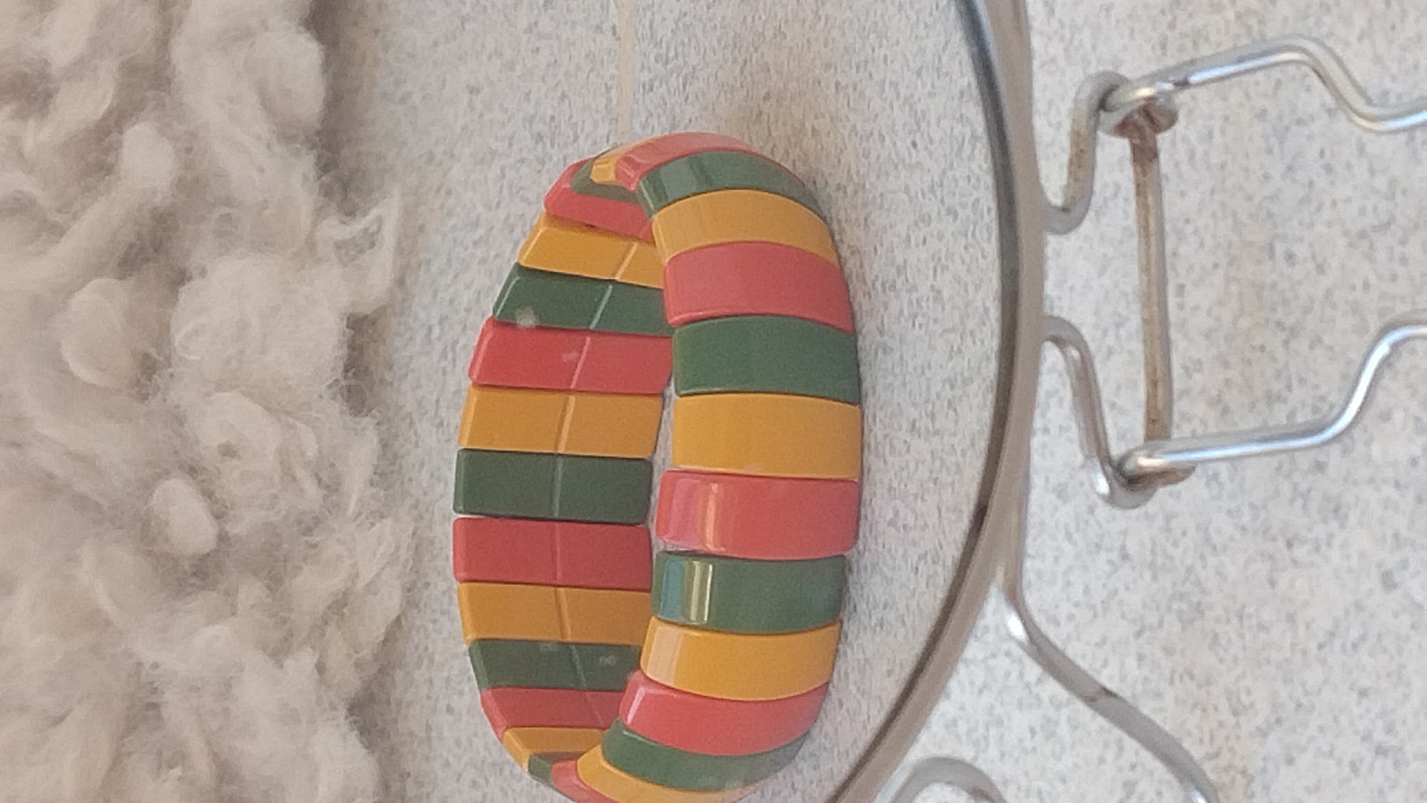 A multi-colored beaded bracelet rests on a speckled gray surface, possibly a countertop or floor. The bracelet is composed of alternating rectangular beads in yellow, red, and green. In the background, a fluffy, off-white material, possibly fabric or cotton, is softly blurred. A portion of a metallic object, appearing to be a rack or holder with curved wires, is visible in the foreground, partially obscuring the bracelet. The overall lighting suggests an indoor setting. There is no information in the image that indicates the location in Perea, Greece.