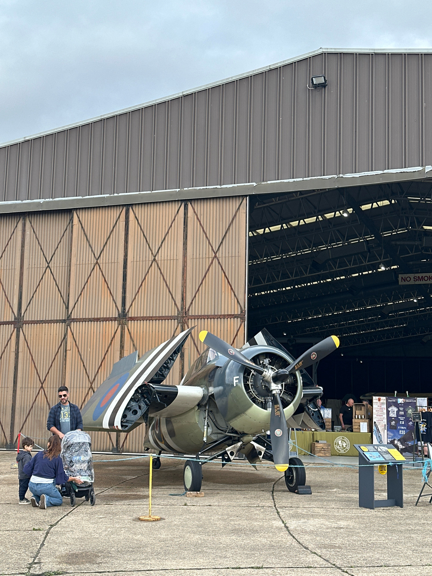 This is a medium shot, eye-level photograph of a World War II-era aircraft, likely a Grumman F4F Wildcat, parked on a concrete tarmac in front of a large, corrugated metal hangar. The aircraft is painted in a camouflage green and brown scheme with RAF roundels on its wings. The hangar doors are open, revealing the dark interior with visible roof trusses and lighting. A "NO SMOKING" sign is visible on the right side of the hangar.
In the foreground, a family is interacting with the aircraft. A man wearing sunglasses and a dark plaid shirt stands behind a woman who is kneeling beside a stroller covered with a clear plastic rain protector. A young child in a grey hooded jacket stands next to the woman. The family appears to be observing or posing with the aircraft.
To the right of the aircraft, a display table with various items, including what appears to be merchandise or information about the aircraft, is set up. A dark grey stand with informational plaques is also present. The overall environment suggests an air show or aviation museum event. The sky is overcast and grey. The scene is set outdoors, on an airfield, possibly within the grounds of the Imperial War Museum Duxford, a well-known aviation museum located near Cambridge, United Kingdom, which frequently displays historical aircraft and hosts events of this nature.