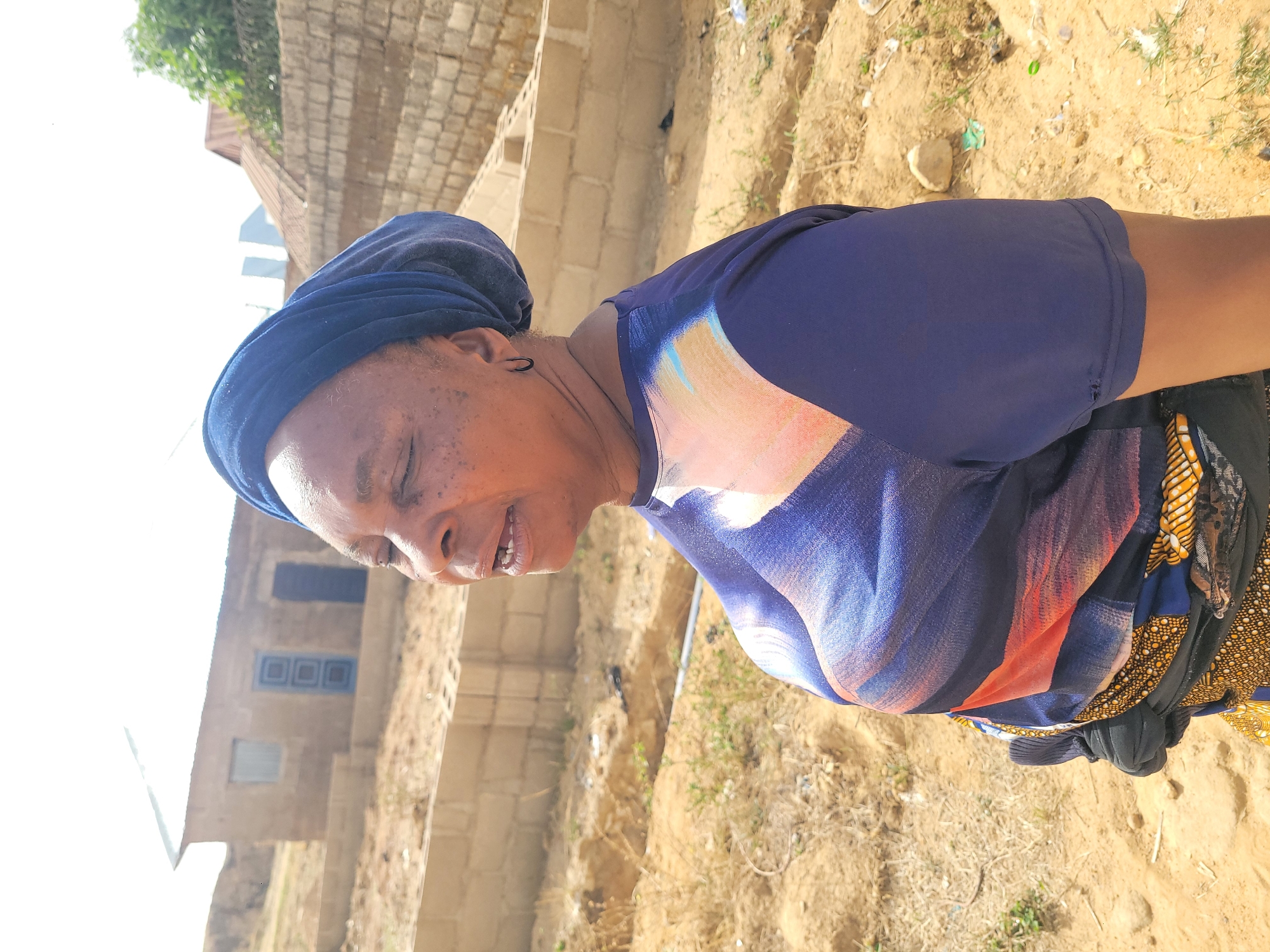 An outdoor, side-profile shot captures a woman smiling in what appears to be a sunny, rural or semi-urban environment in Barakin Akawo, Nigeria. The woman, with a dark complexion, is seen from the chest up, looking towards the left side of the frame. She wears a dark blue head wrap or scarf and a short-sleeved top featuring a vibrant abstract pattern with shades of blue, orange, and black. A small, dark hoop earring is visible in her left ear. Her eyes are partially closed in a joyful expression, and her mouth is slightly open, indicating a light smile or laugh.
The background consists of sandy, reddish-brown dirt ground with sparse patches of dry grass and small stones. Behind the woman, several structures built with unplastered brick or concrete blocks are visible, suggesting unfinished construction or older buildings. One building features three square blue window frames or decorative elements. The left side of the image is significantly overexposed by a bright light source, likely the sun, indicating clear, sunny daytime conditions. No text is visible in the image.