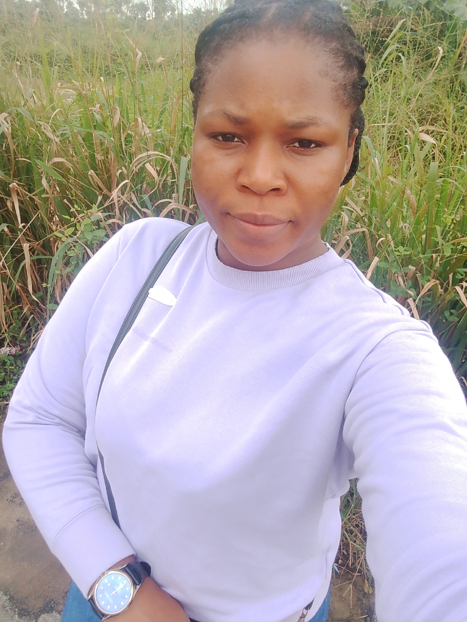 In Yenegoa, Nigeria, a woman with braided hair takes a selfie outdoors. She is wearing a light purple sweatshirt and blue jeans. A dark-strapped watch is visible on her left wrist. She is standing in front of tall, dry grass. A dark-colored strap from a bag is visible across her chest. The lighting suggests it is daytime.