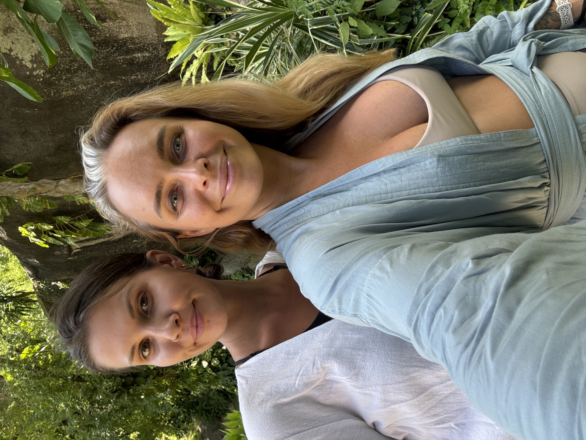 Two women, one with dark hair and a nose ring, and another with blonde hair and grey streaks, pose for a selfie outdoors. They are surrounded by lush green foliage, suggesting a tropical environment. The woman on the left wears a white garment over a black bra, while the woman on the right is in a light blue robe over a beige bra. The background includes a rocky wall and dense vegetation, characteristic of a natural setting in Ko Samui District, Thailand. The lighting indicates it is daytime.