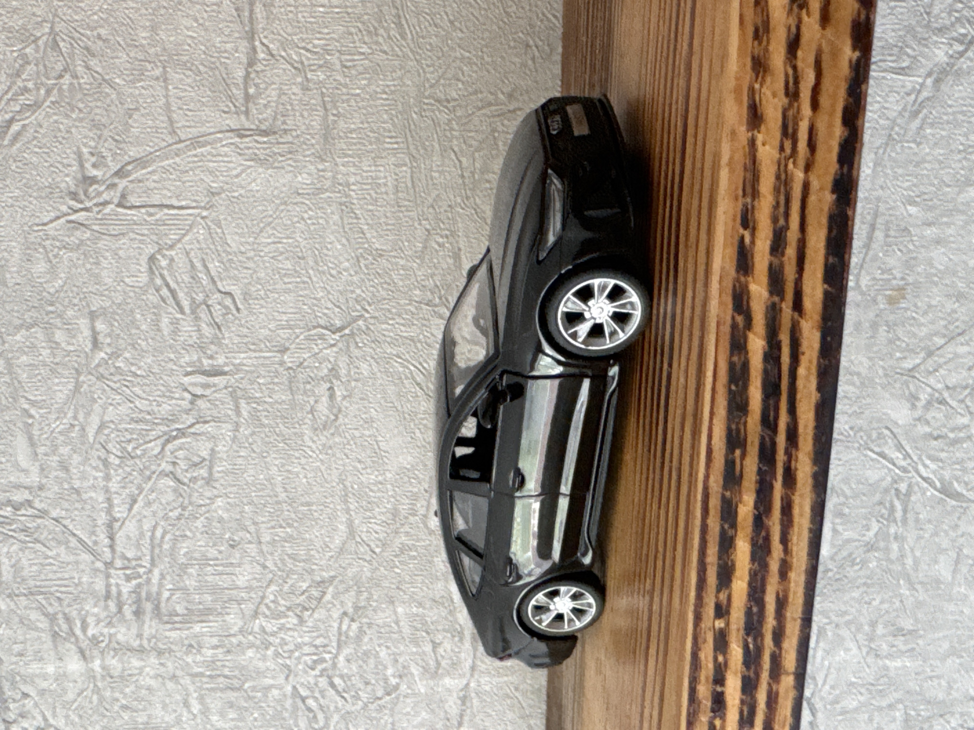 A black toy Audi A7 is positioned on a wooden surface. The car is viewed from a slightly elevated side-angle, showcasing its sleek design. The background consists of a textured, off-white wall. The image is taken in what is described as the city of Яромінскі сельскі Савет, Belarus, although no specific urban or geographical features are visible in the frame to corroborate this location. The lighting is neutral, and the overall scene is static, with no visible human presence or activity.