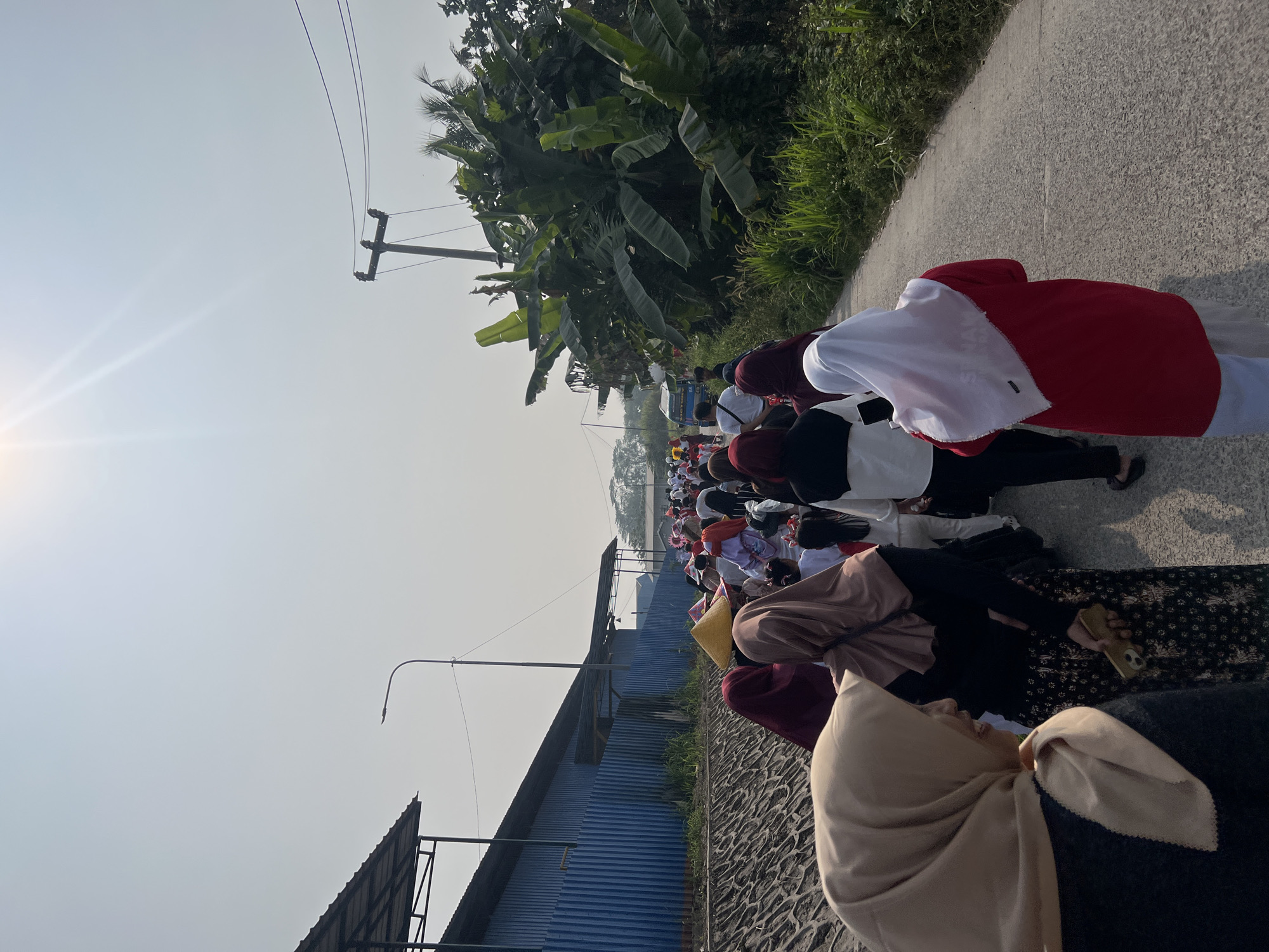 This is a horizontal, medium shot photograph taken from a slightly low angle, looking down a rural road in Klapanunggal, Indonesia. The sky is bright and hazy, with the sun radiating from the top center of the frame. A large crowd of people, predominantly women wearing hijabs and long dresses, are walking away from the camera down the road. Many are wearing white or red clothing, and some have conical hats. The road is paved with asphalt and is lined on the left by a low, stone wall and a large, blue industrial building with a corrugated metal roof. On the right side of the road is a dense growth of vegetation, including large-leafed plants that appear to be banana trees, and tall grasses. A utility pole with wires stands along the right side of the road, and a small blue van is visible in the distance. The overall impression is a religious or cultural procession taking place on a sunny day.