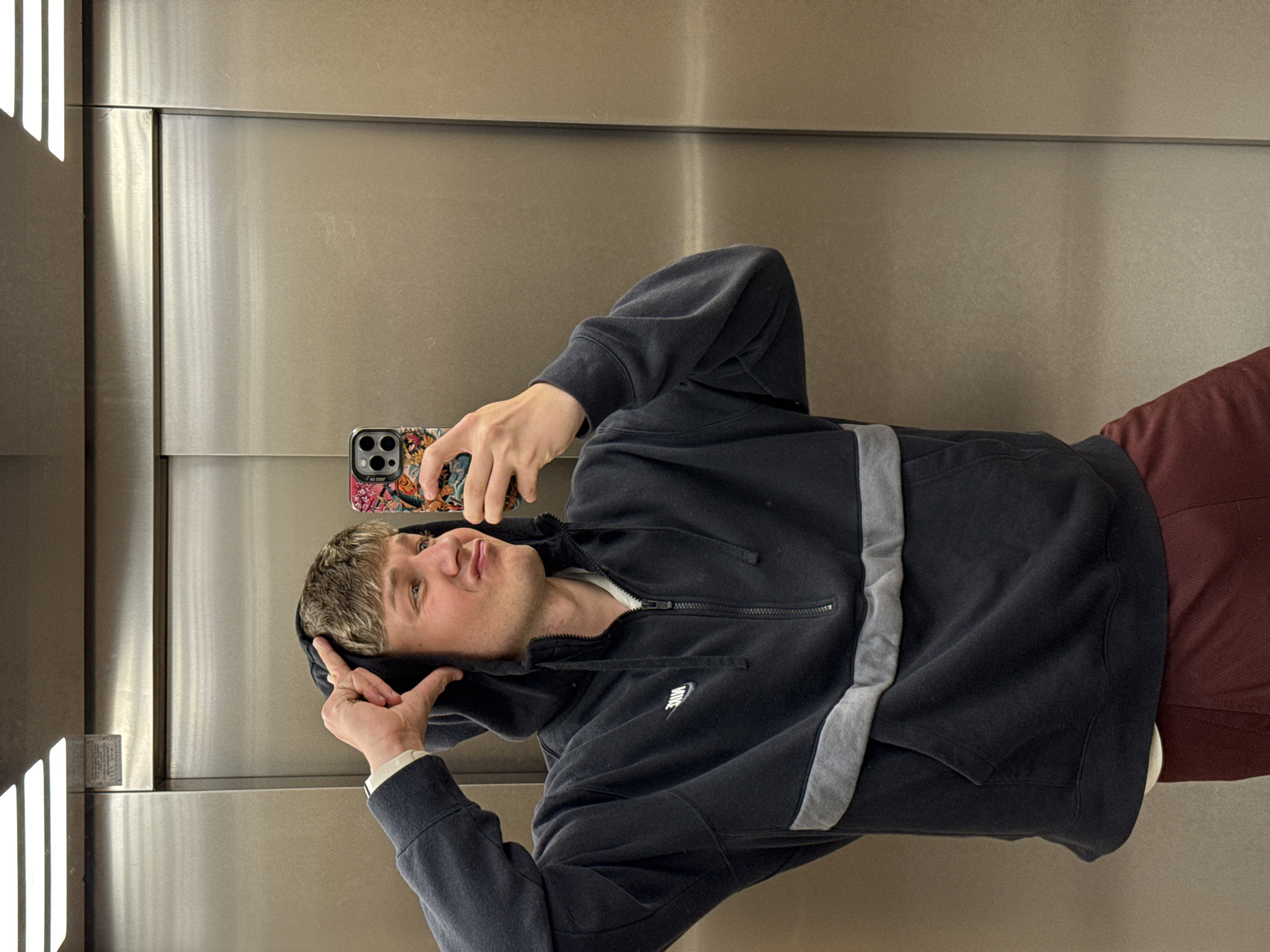 A young man is taking a selfie in what appears to be an elevator. He is wearing a dark gray Nike hoodie with a gray stripe down the side, and burgundy pants. He is holding a phone with a colorful case. The background is the elevator walls, which are made of a brushed metal material. The location is not discernible as Plovdiv, Bulgaria, or anywhere else specifically from the image.