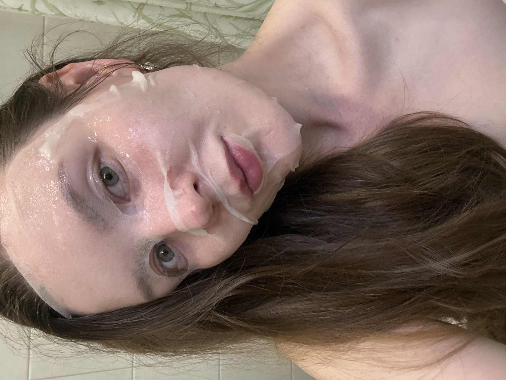 A woman with light skin is wearing a sheet mask on her face. Her eyes are green, and her hair is brown. She appears to be lying down, and the photo is taken from a slightly above perspective. The background shows a tiled surface, likely a bathroom. There is no indication of the city Izhevsk, Russia in the image.