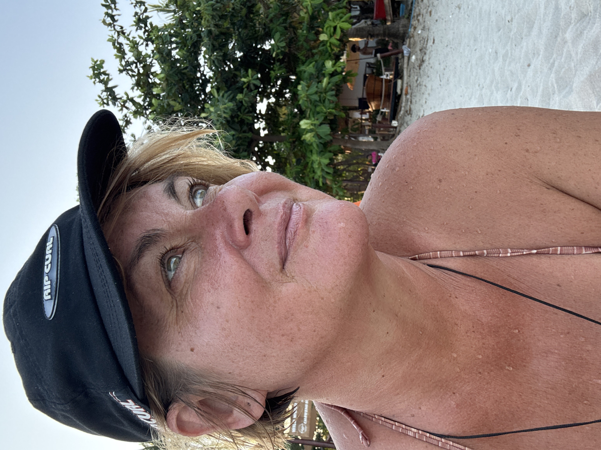 A medium shot of a woman wearing a black Rip Curl baseball cap, a bikini and standing on a sandy beach in Baan Chaweng, Thailand. She is looking upwards. The background features tropical trees and a beach bar or refreshment stand. The woman's skin is damp.