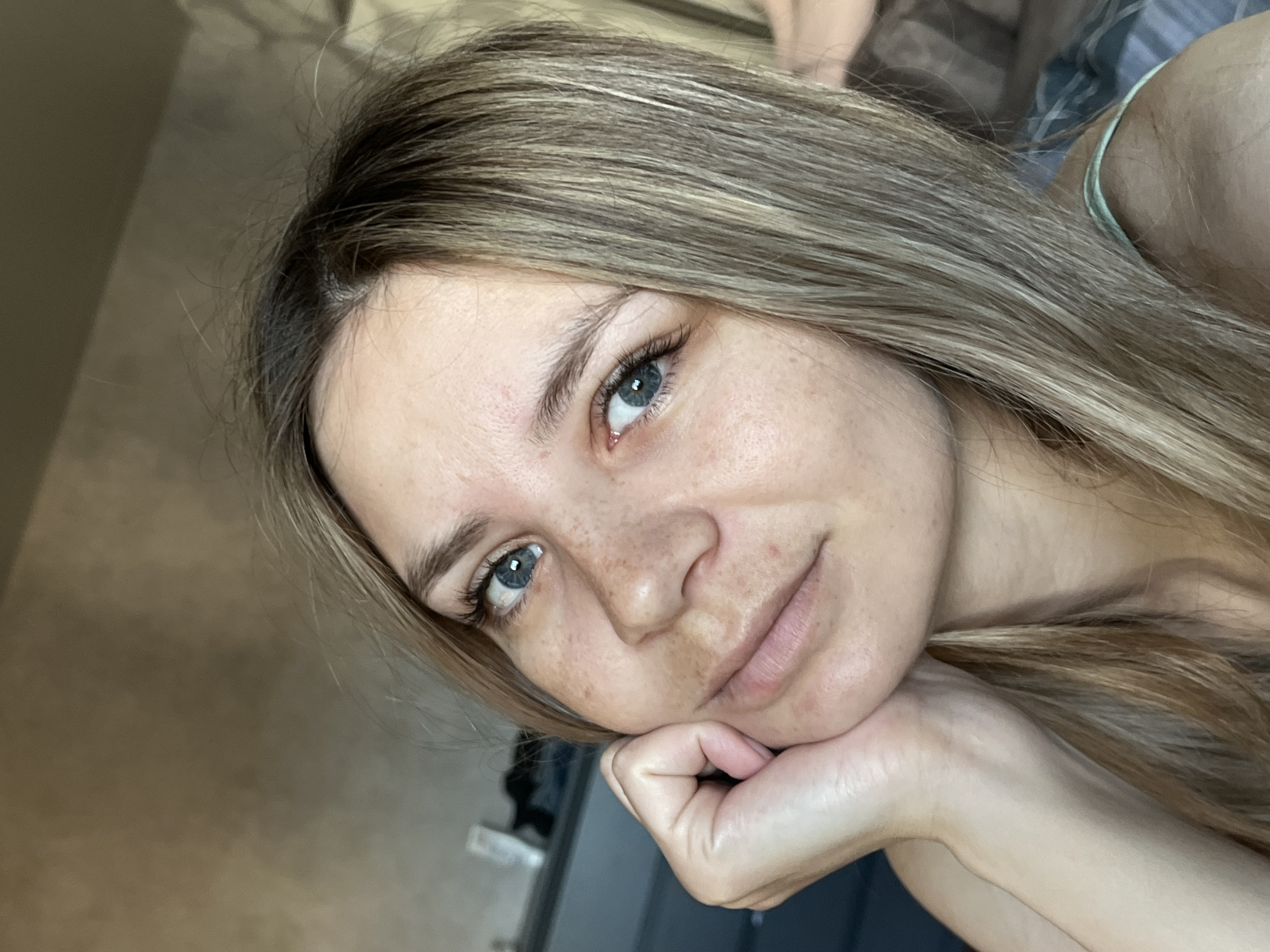A young woman with light brown hair and blue eyes is posing for a close-up selfie. She is resting her chin on her left hand, looking slightly upwards with a subtle smile. The background appears to be an indoor setting with a wall visible on the left and a glimpse of clothing and furniture behind her. The image was taken indoors, location, if any, cannot be determined.