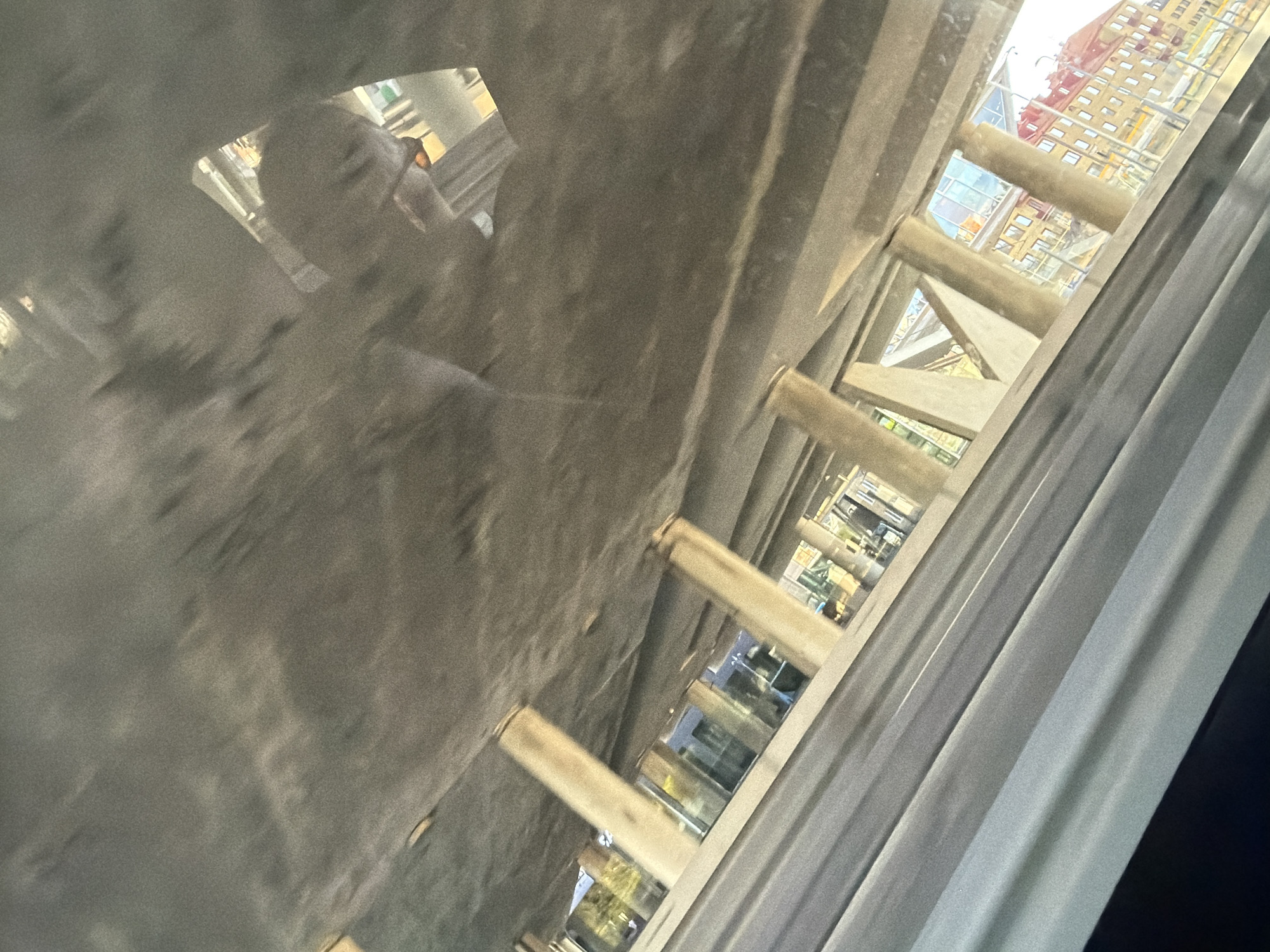 The image shows a view from under a concrete structure, possibly a bridge or overpass, looking out towards buildings and a road. Several large concrete pillars support the overhead structure. Through the gaps between these pillars, a street with cars can be seen, along with buildings featuring red and yellow facades. A reflection of a person's face is visible on the underside of the concrete structure. The scene appears to be a busy urban or suburban area.