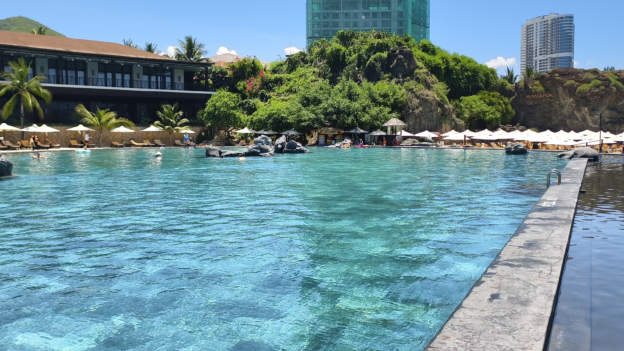 The image shows a large outdoor swimming pool with crystal-clear turquoise water. In the background, there is a resort with a long, brown-tiled building and lush green vegetation on a rocky hillside. Several white umbrellas and lounge chairs are set up along the edge of the pool. People are seen swimming and relaxing in and around the pool. In the distance, there are tall buildings, indicating an urban setting. The sky is clear and blue. The scene appears to be a sunny day at a resort.