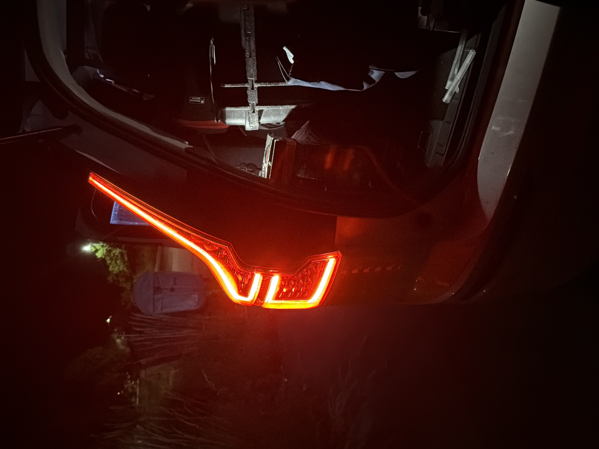 The image depicts the rear of a car with its trunk open, illuminated by the car's red taillights and some ambient light from the surroundings. The scene appears to be outdoors, possibly a parking lot or roadside, with some vegetation and a light post visible in the background. The trunk contains various objects, including what appear to be bags and possibly some equipment. The overall lighting suggests it is nighttime or dusk. The image does not contain any identifiable landmarks or clues that would place it specifically in Smyadovo, Bulgaria.